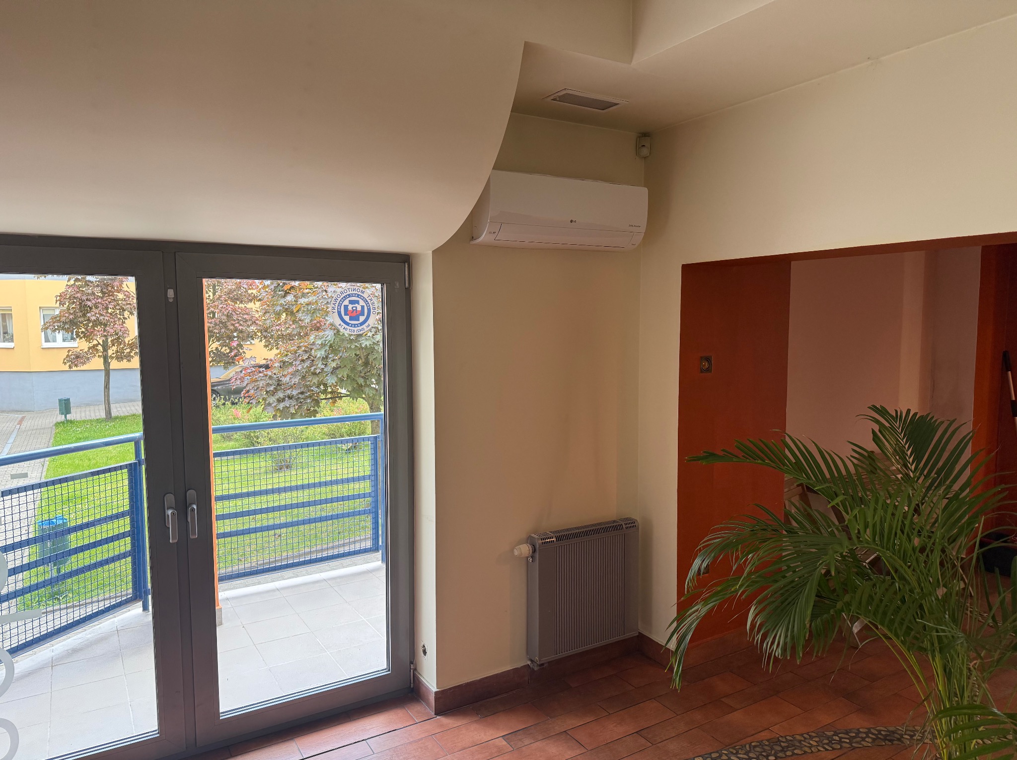 Biała klimatyzacja LG na ścianie w jasnym wnętrzu z oknem balkonowym i grzejnikiem. Widoczna zieleń za oknem i roślina doniczkowa w pomieszczeniu.