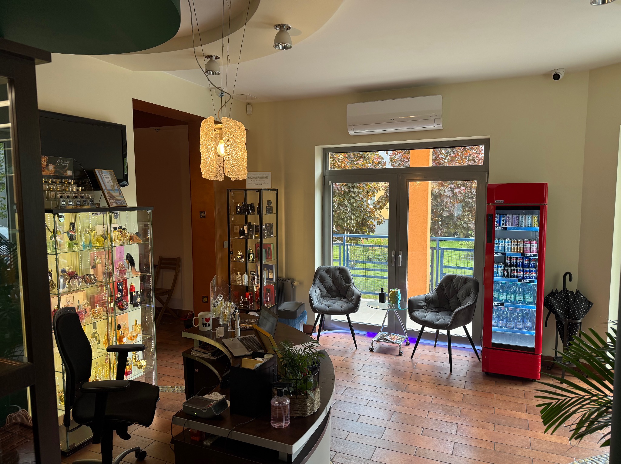 Wnętrze biura z klimatyzacją LG nad drzwiami balkonowymi, szklana witryna z perfumami, dwa fotele i czerwona lodówka z napojami.