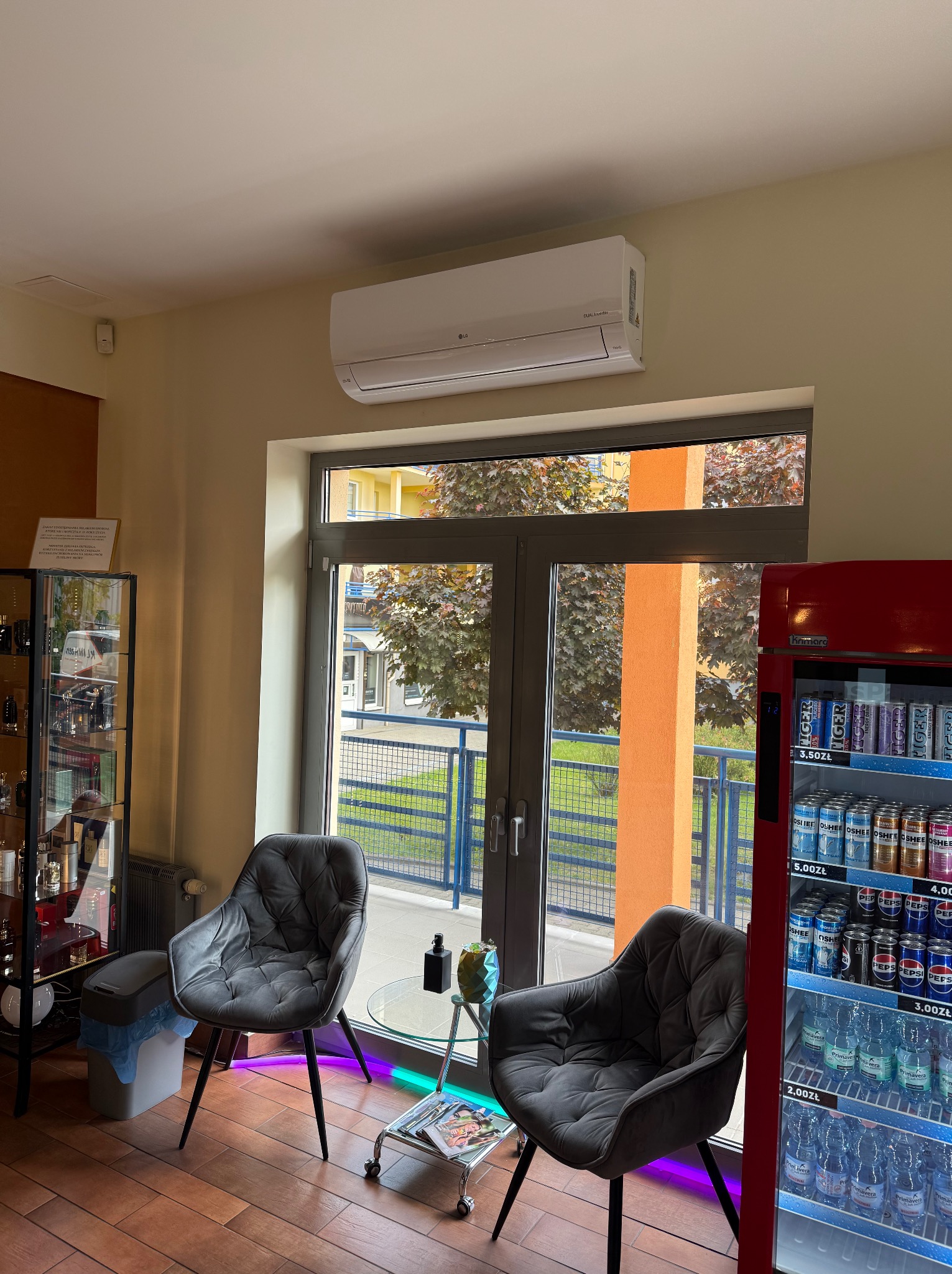 Wnętrze lokalu z klimatyzatorem LG Dual Inverter nad oknem. Widoczne krzesła, stolik kawowy i lodówka z napojami. Na podłodze płytki imitujące drewno.