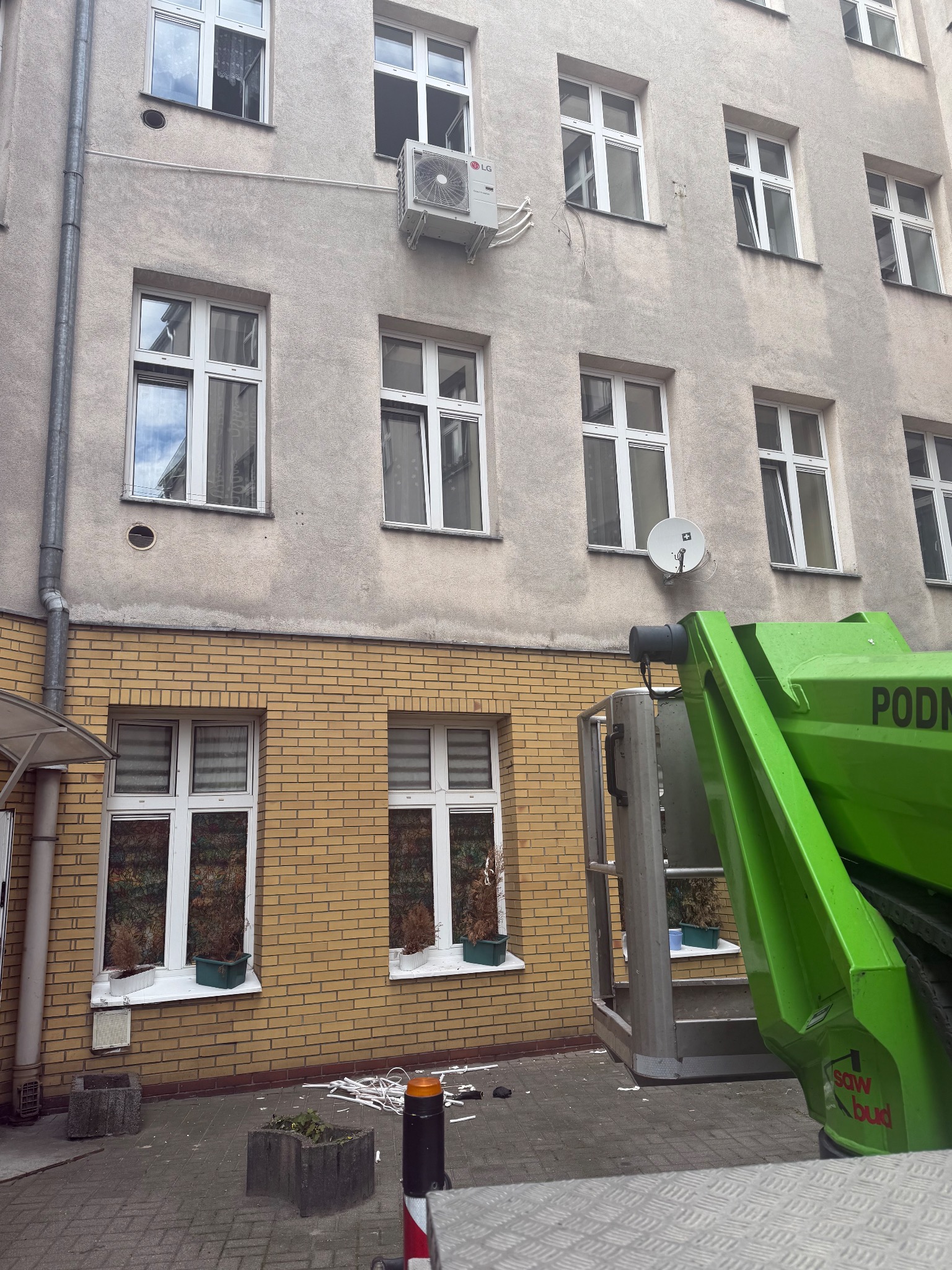 Szara elewacja budynku z oknami i zamontowaną klimatyzacją LG, widok z podnośnika koszowego podczas prac instalacyjnych. Kabel na chodniku.