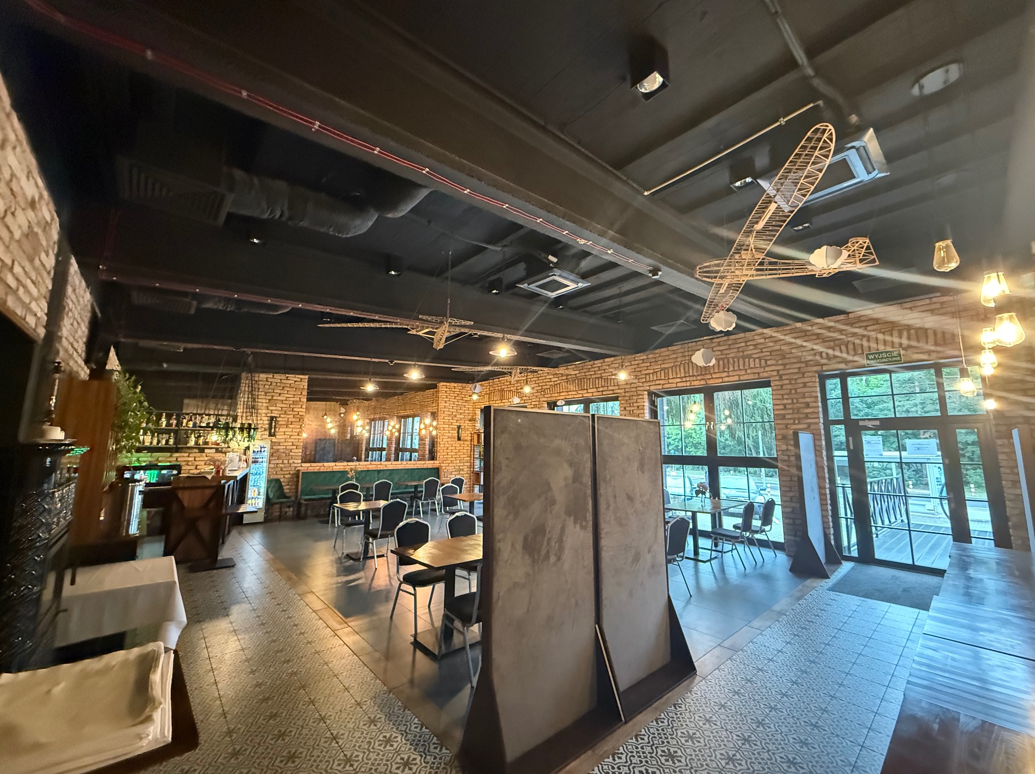 Wnętrze restauracji z widocznymi elementami wentylacji pod sufitem, dekoracyjne modele samolotów i ceglane ściany. Widoczne kratki wentylacyjne.