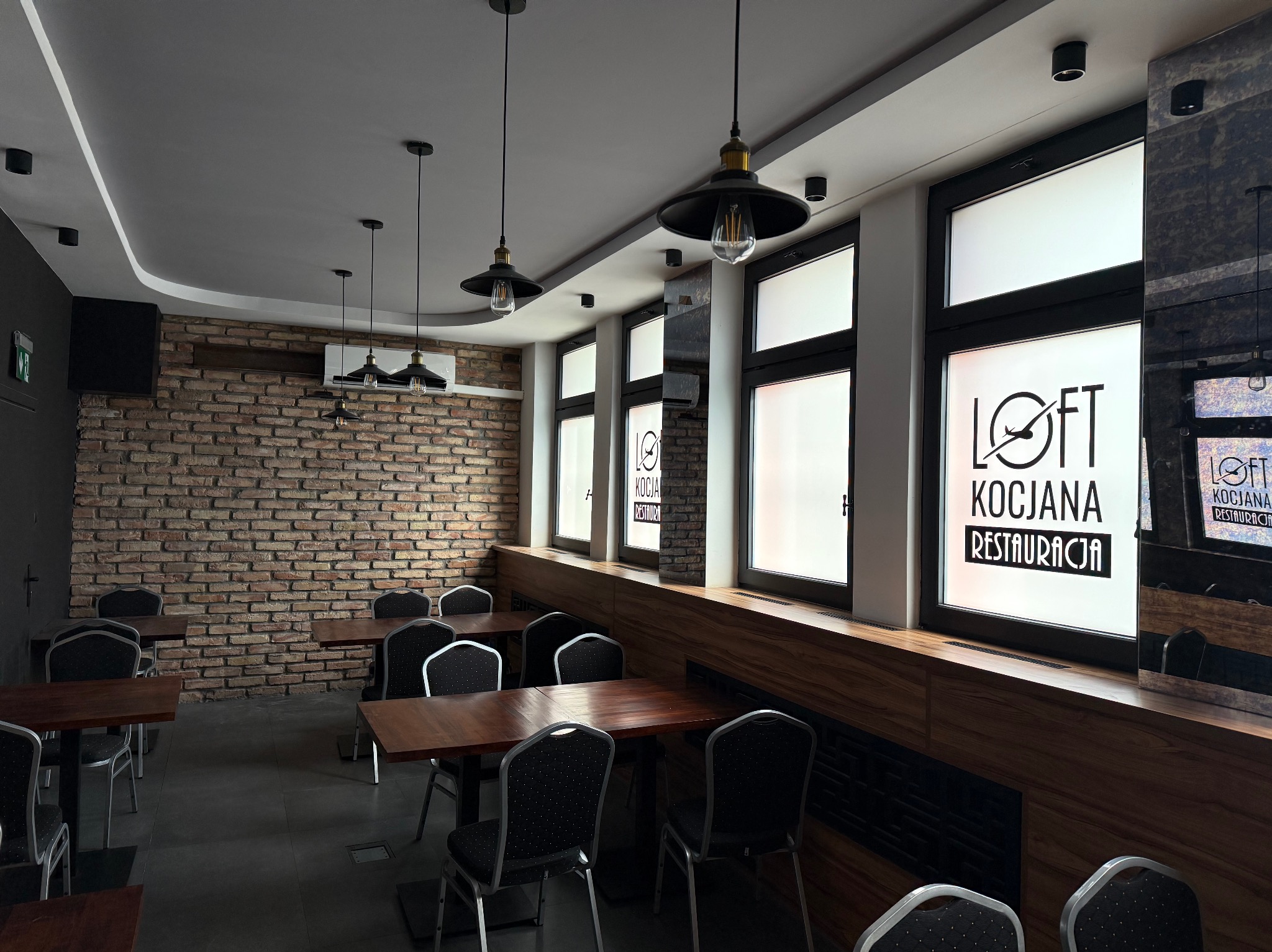 Wnętrze restauracji w stylu loft z ceglaną ścianą, wiszącymi lampami i klimatyzacją. Widoczne stoły i krzesła. Na oknach logo restauracji.