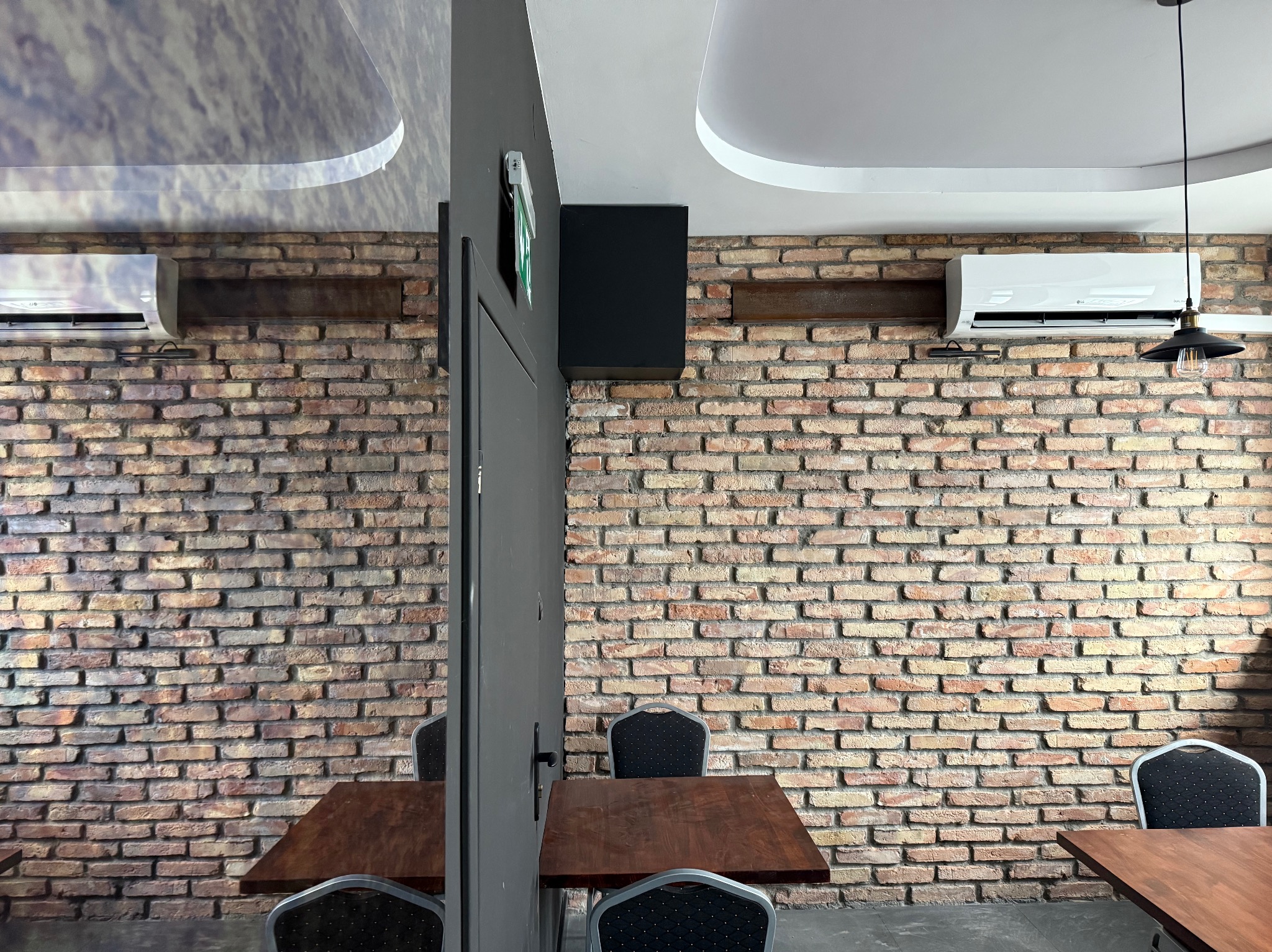 Klimatyzator LG zamontowany na ceglanej ścianie w lokalu. Widoczny stół i krzesła. Na suficie lampa i element dekoracyjny. Część klimatyzatora odbija się w lustrze.