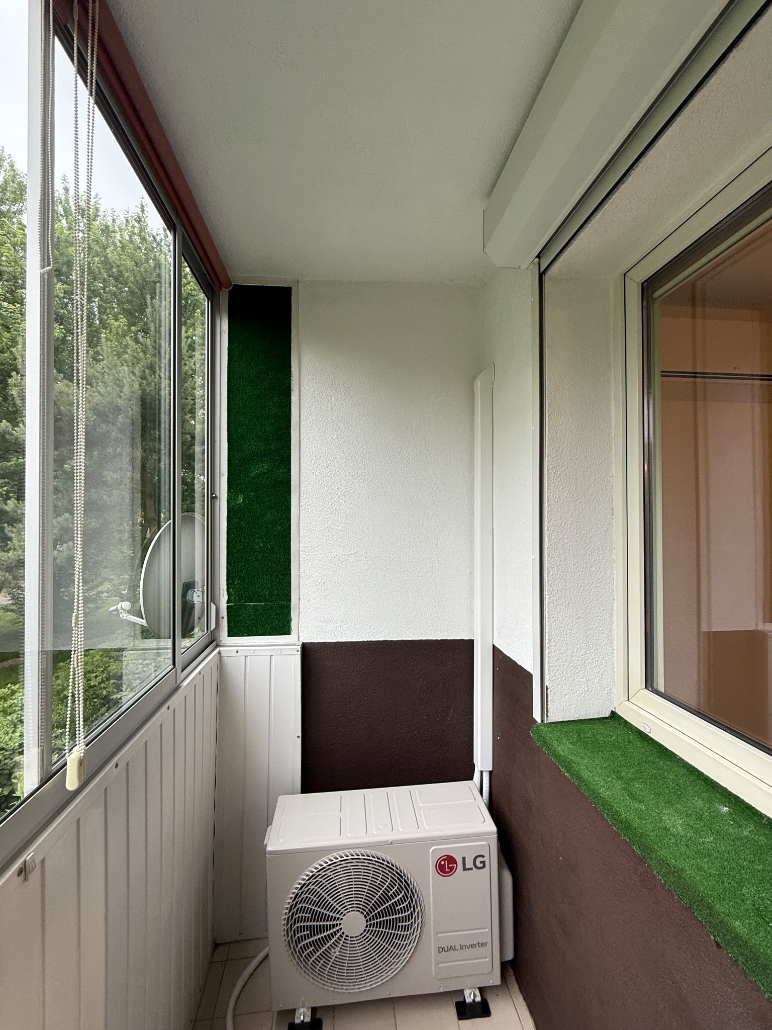 Biała klimatyzacja LG Dual Inverter na balkonie z panelami PCV, sztuczną trawą i dużymi oknami. Widok na zieleń z zewnątrz.