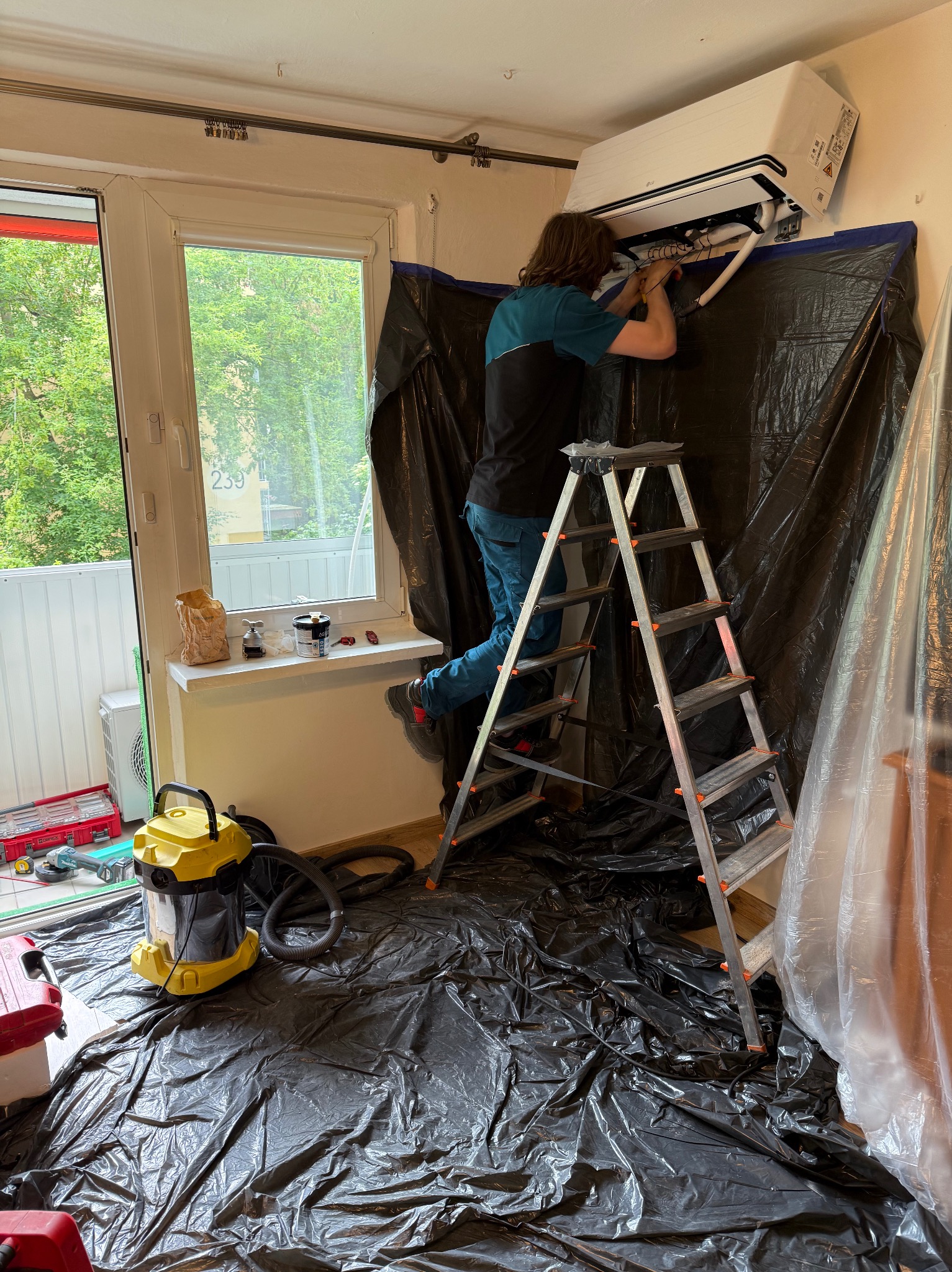 Instalacja klimatyzacji w Łodzi: mężczyzna na drabinie montuje jednostkę ścienną, zabezpieczone folią, widoczny odkurzacz i narzędzia.