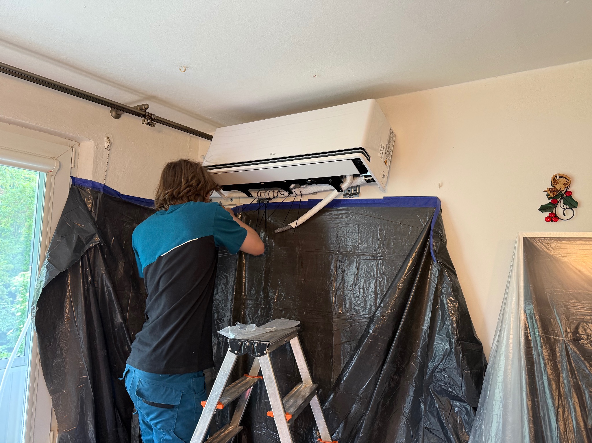 Instalator montuje klimatyzator LG w pomieszczeniu zabezpieczonym folią ochronną. Widoczna drabina i przewody. Biały klimatyzator na jasnej ścianie.