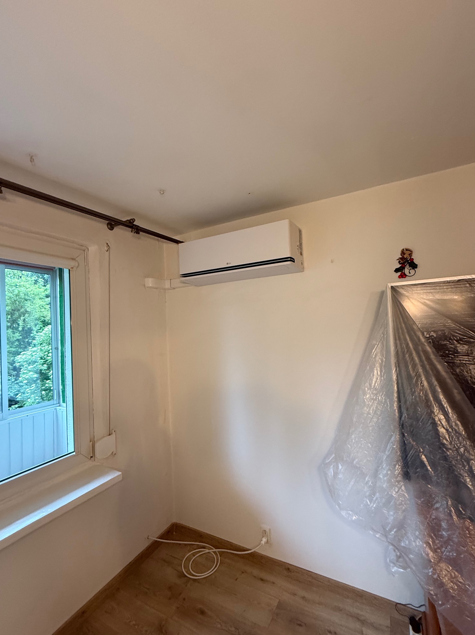 Zamontowana, biała klimatyzacja LG w jasnym pokoju z oknem i zasłoniętym meblem. Widoczny kabel zasilający.