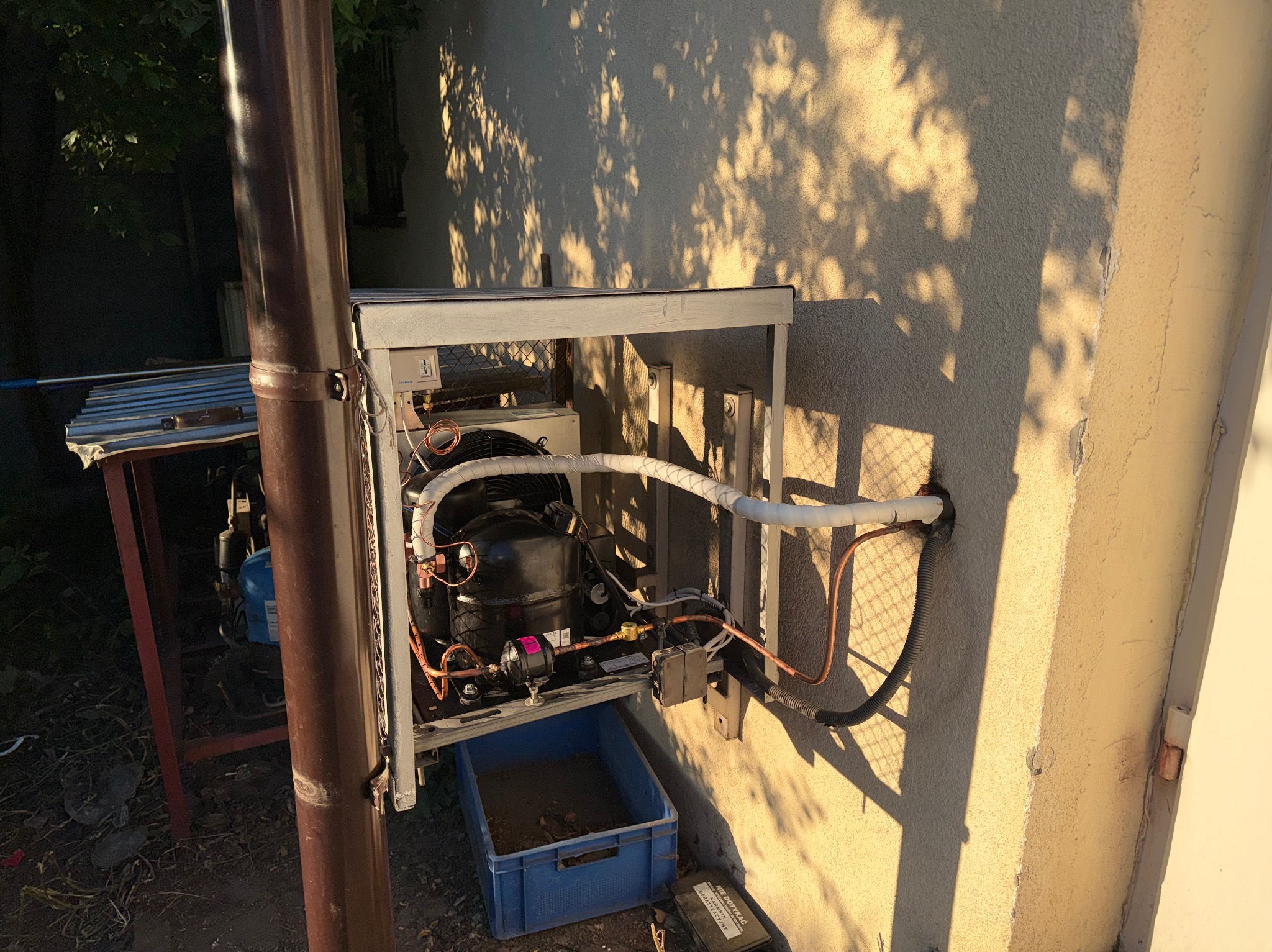 Zewnętrzna jednostka klimatyzacji z widocznymi rurami miedzianymi i elementami instalacji, zamontowana na ścianie budynku w słoneczny dzień. Widoczne cienie liści na elewacji.