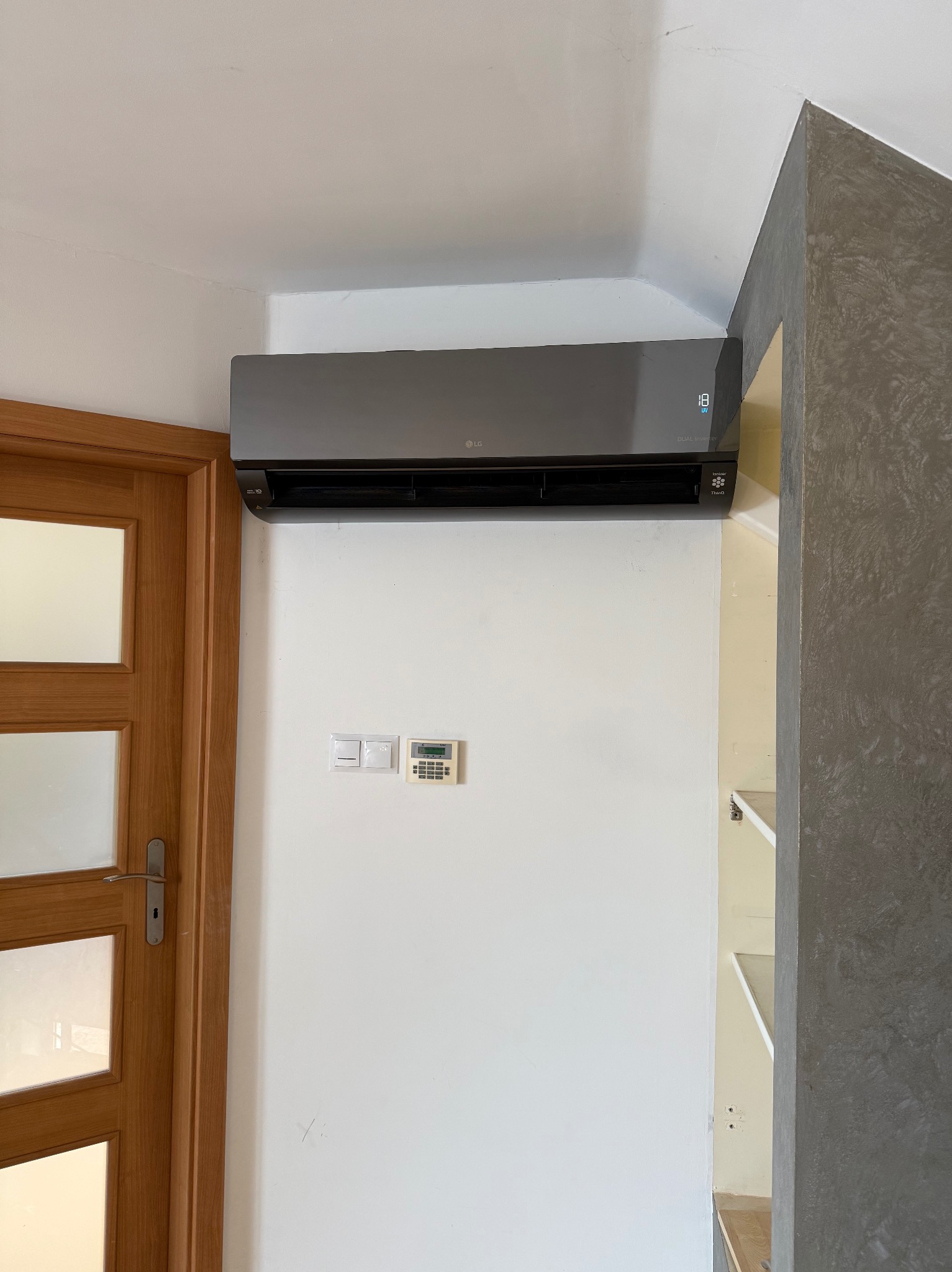 Czarny klimatyzator LG Dual Inverter zamontowany na białej ścianie w pomieszczeniu. Widoczne panele sterowania i fragment drewnianych drzwi z przeszkleniami.