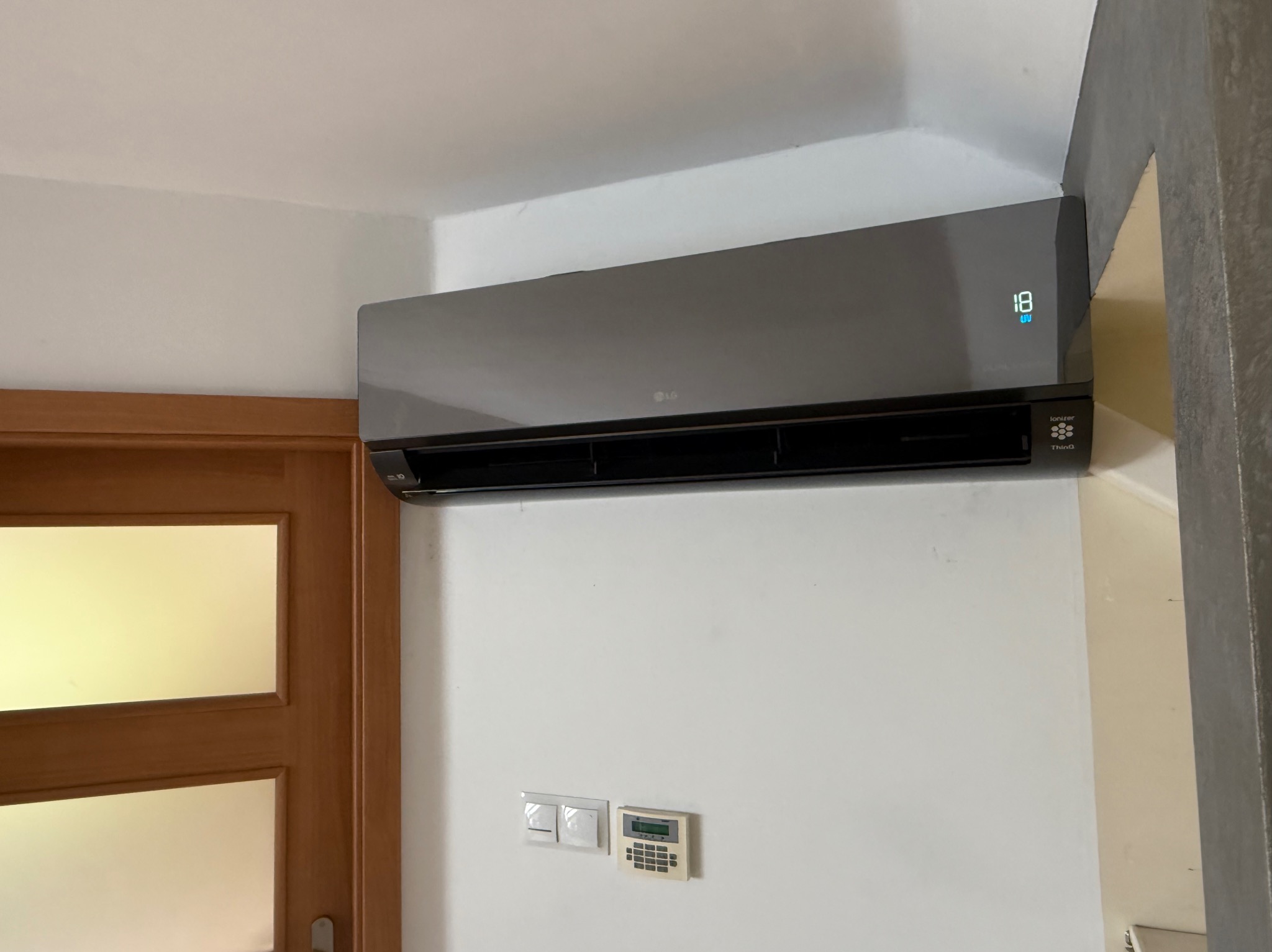 Szary klimatyzator LG, zamontowany na białej ścianie, obok drewnianych drzwi. Widoczny panel sterowania i wyświetlacz temperatury. Nowoczesny design.