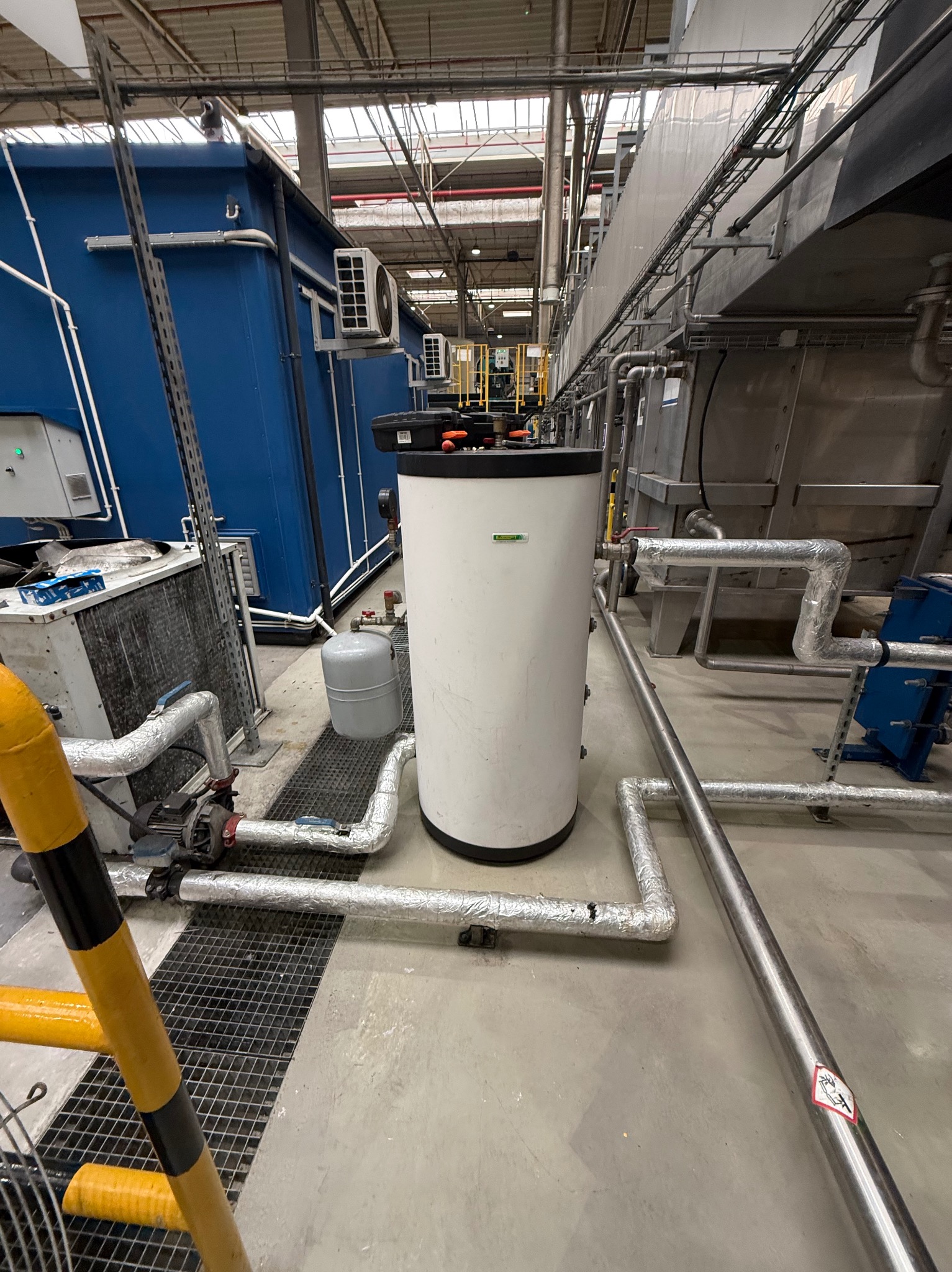 Biały zbiornik buforowy z czarną górą w przemysłowym wnętrzu. Widoczne rury, instalacje, klimatyzatory na ścianie i drabina z żółto-czarnymi oznaczeniami.