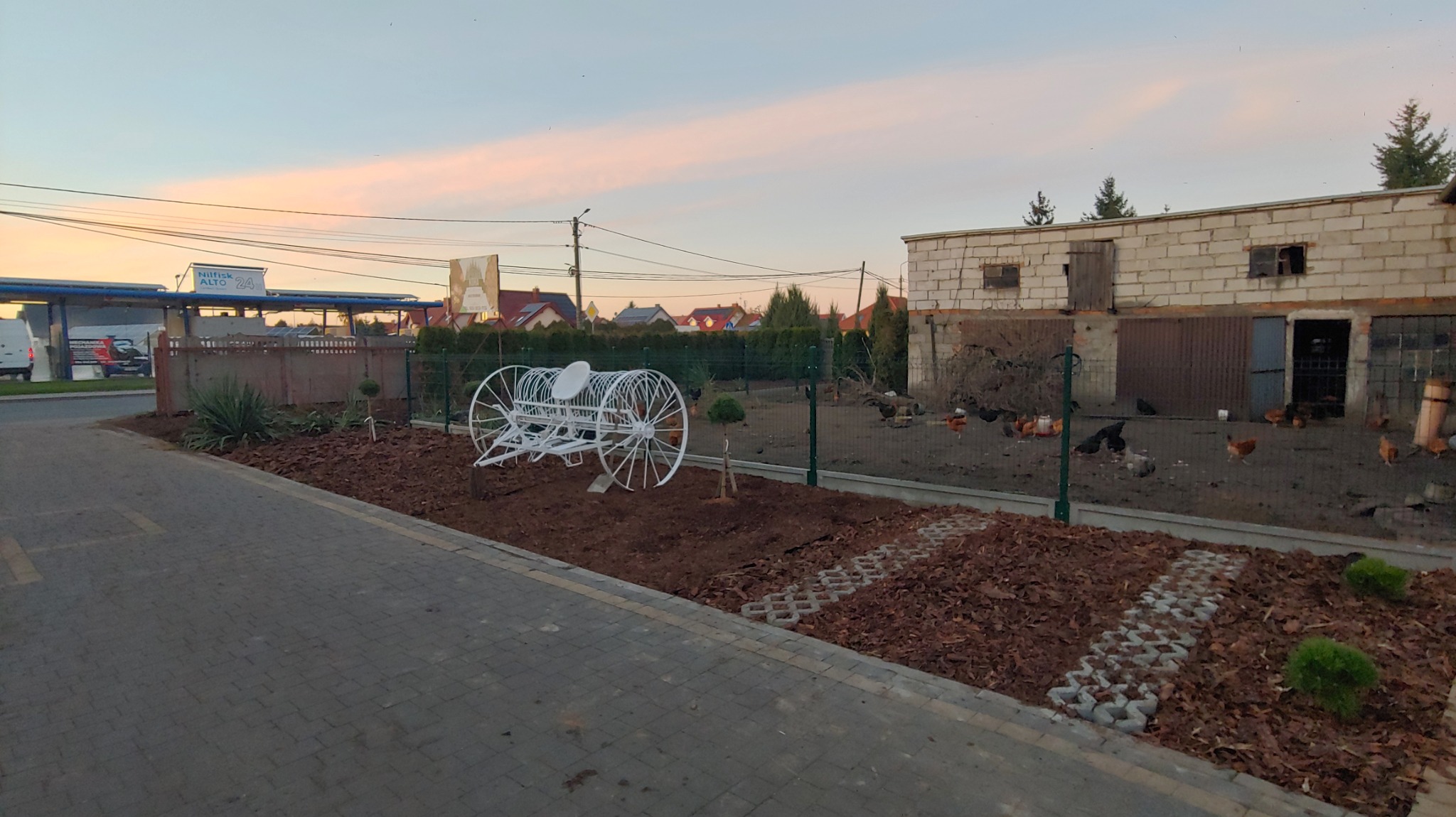 Nowo założony ogród z dekoracyjną, białą konstrukcją w kształcie wozu, wyściółkowany korą, z młodymi drzewkami i kostką ażurową. W tle budynek gospodarczy i zabudowa jednorodzinna.