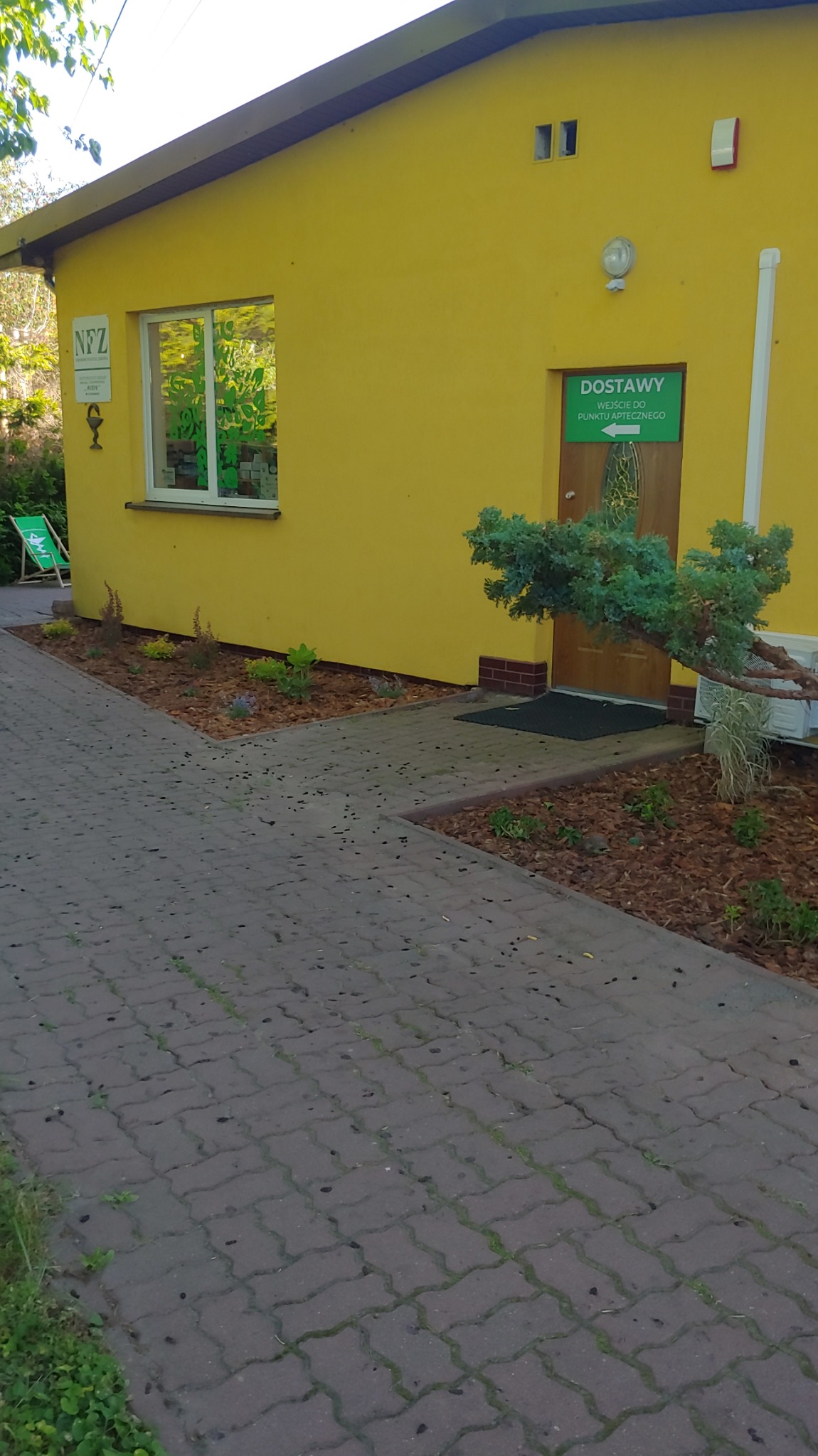 Żółty budynek z nowymi nasadzeniami roślin i korą wokół, ścieżka z kostki brukowej prowadzi do wejścia. Dekoracyjny krzew iglasty przy drzwiach.