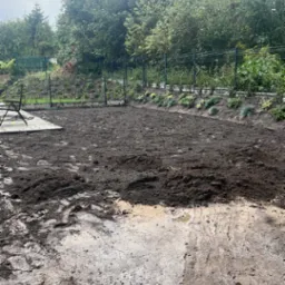 Przygotowany teren pod trawnik, świeżo przekopana ziemia z widocznymi śladami opon, w tle ogrodzenie z siatki i linia zasadzonych roślin przy obrzeżu.