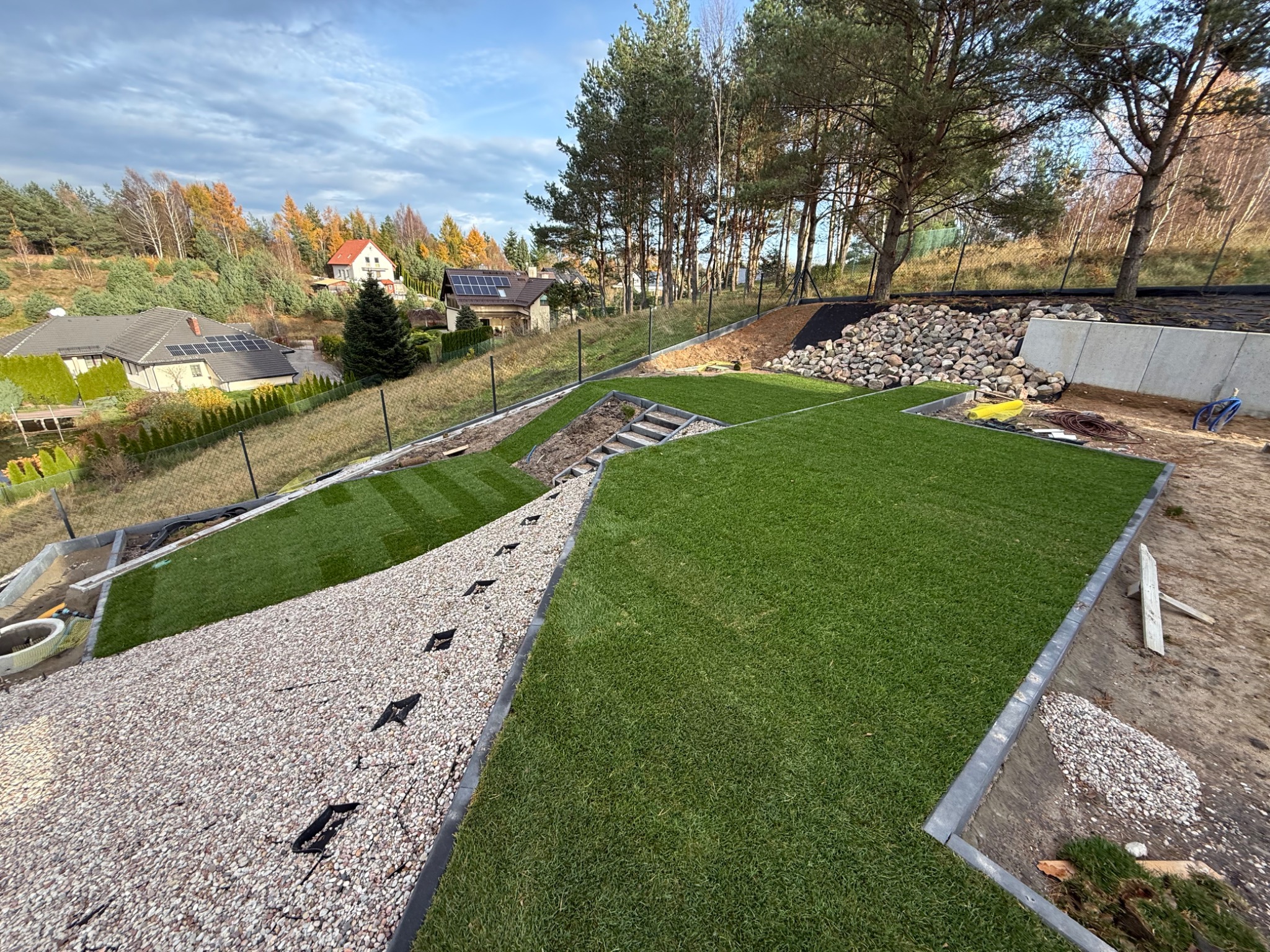 Ukończony ogród tarasowy z równo ułożoną trawą z rolki i ścieżką z jasnego żwiru, otoczony zielenią i domami w tle, w słoneczny dzień.