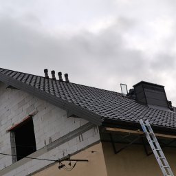 Usługi budowlane WenDach - Dom w trakcie budowy z nowym, ciemnym dachem z blachodachówki, widoczne kominy wentylacyjne, drabina oparta o dach, ściany z bloczków i fragment elewacji w kolorze beżowym.