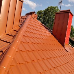 Usługi budowlane WenDach - Nowy, miedziany dach z blachy trapezowej z kominami i anteną na tle błękitnego nieba z chmurami, zieleń drzew w tle. Widok z góry.