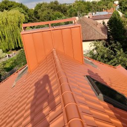 Usługi budowlane WenDach - Nowy, pomarańczowy dach z dachówki ceramicznej, z oknem dachowym i kominem. Cień osoby na dachu. W tle zieleń drzew i zabudowa miejska.