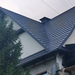 Usługi budowlane WenDach - Szary dach z blachy na dwuspadowym budynku, widok z dołu, z fragmentem drzewa iglastego po lewej stronie. Widoczne okno i fragment rusztowania.