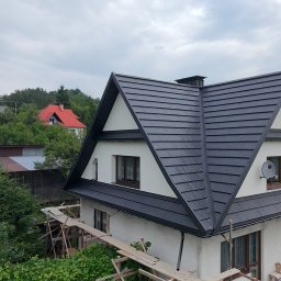 Usługi budowlane WenDach - Dom z czarnym dachem w kształcie litery A, widok z góry, otoczony zielenią. Widoczne rusztowania. W tle wzgórza i inne budynki.