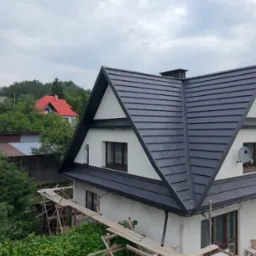 Dom z czarnym dachem w kształcie litery A, widok z góry, otoczony zielenią. Widoczne rusztowania. W tle wzgórza i inne budynki.