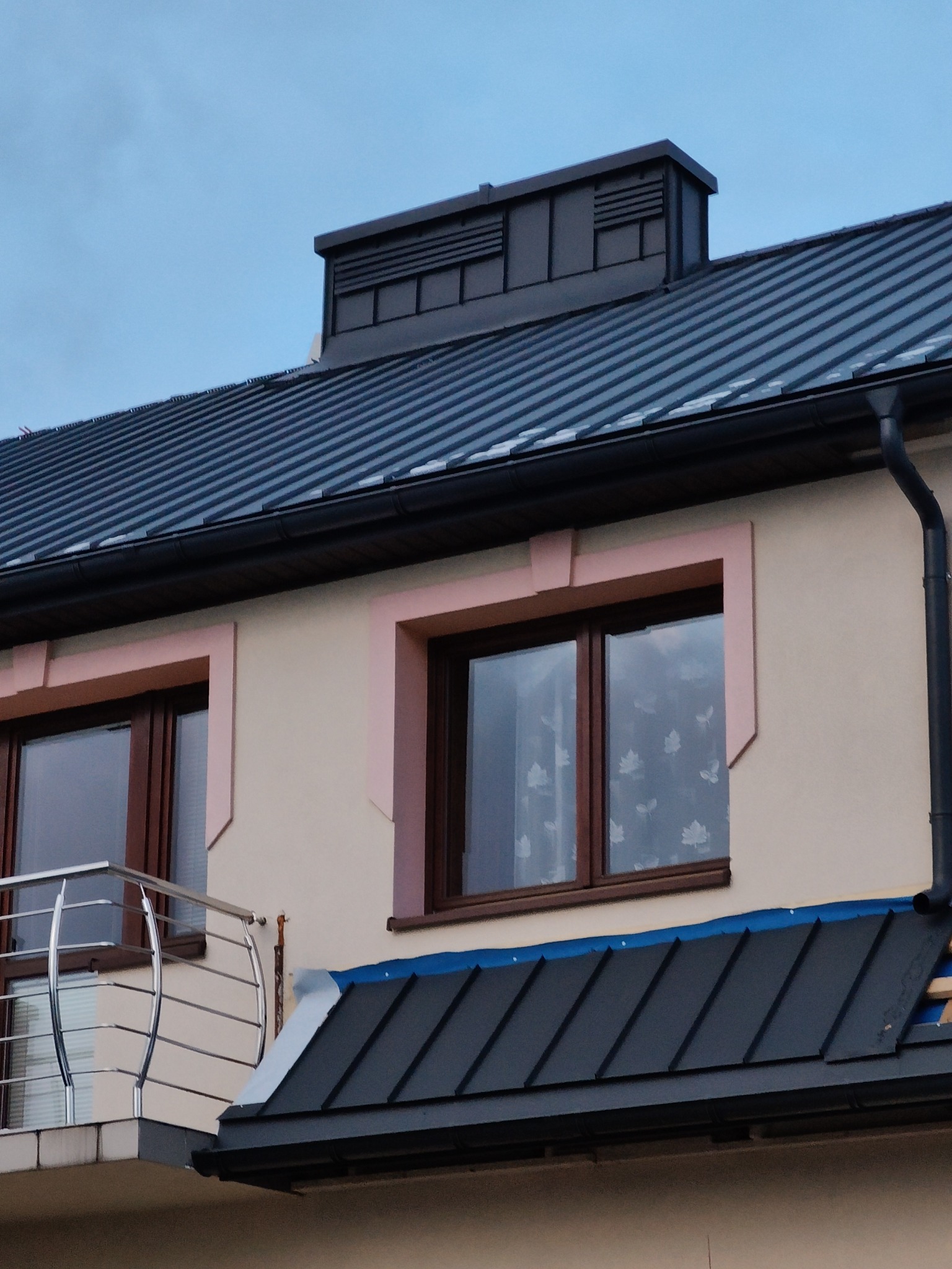 Fragment domu z nowym, ciemnym dachem z blachy trapezowej, widoczny komin wentylacyjny, rynna oraz okna z jasnoróżowymi obramowaniami.