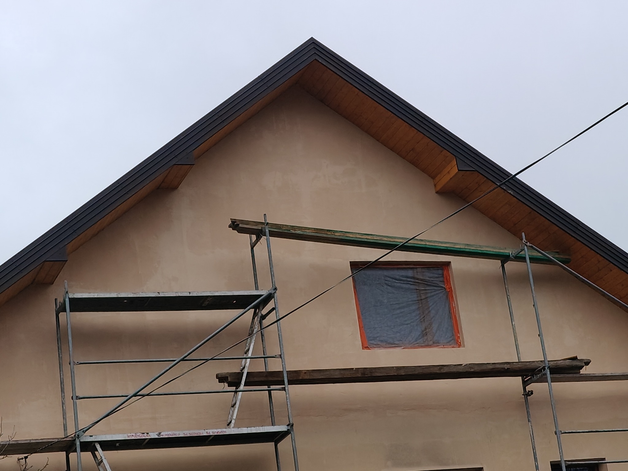 Dom w trakcie prac wykończeniowych elewacji, widoczne rusztowania i zabezpieczone okno folią, nowy dach z ciemnej blachy trapezowej z drewnianą podbitką.