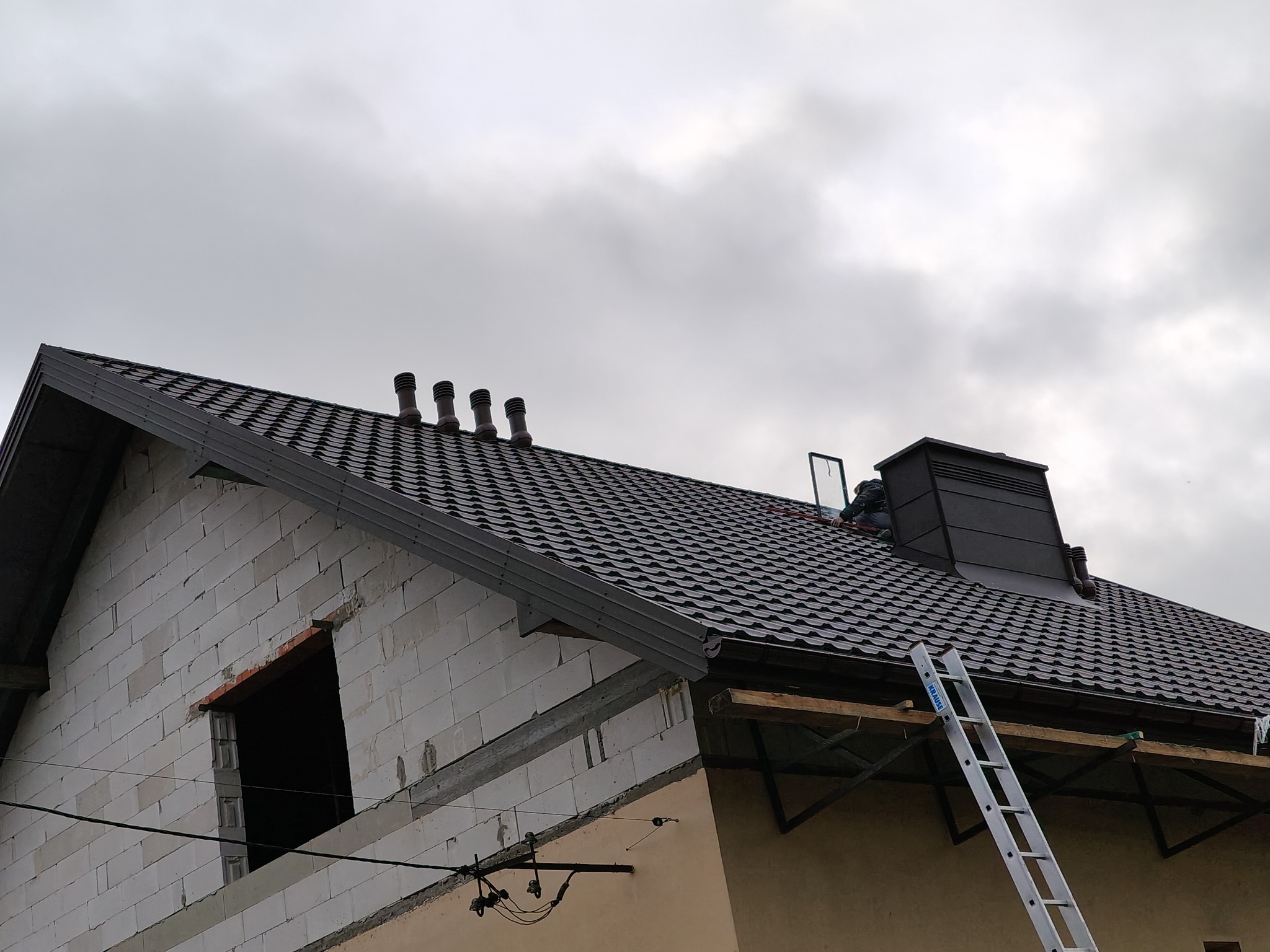 Dom w trakcie budowy z nowym, ciemnym dachem z blachodachówki, widoczne kominy wentylacyjne, drabina oparta o dach, ściany z bloczków i fragment elewacji w kolorze beżowym.