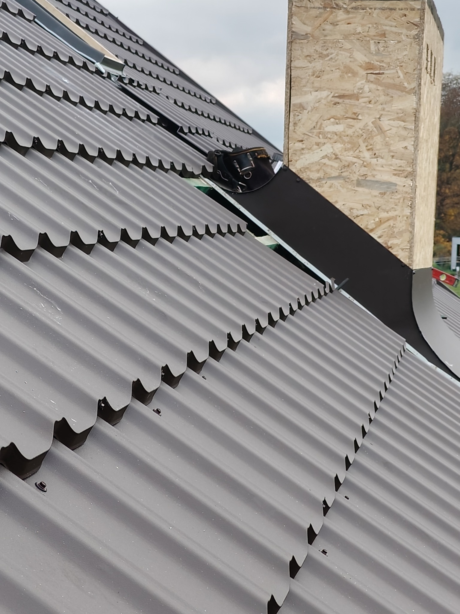 Nowy, brązowy dach z blachy trapezowej, z widocznym kominem w trakcie obróbki blacharskiej i narzędziami dekarskimi, na tle pochmurnego nieba.