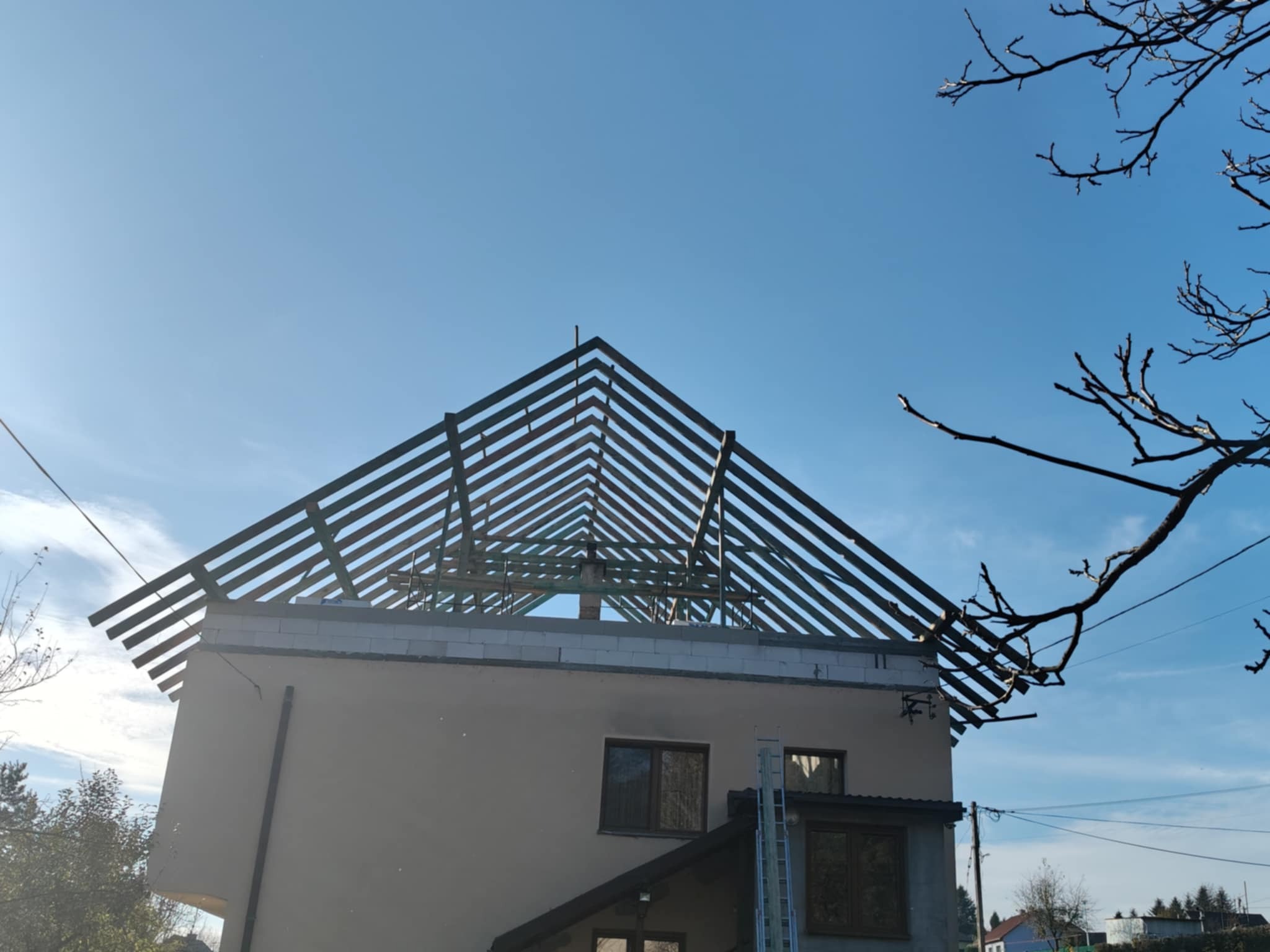 Dom jednorodzinny w trakcie budowy dachu, widoczna drewniana konstrukcja więźby dachowej na tle błękitnego nieba, z fragmentem drzewa z prawej strony