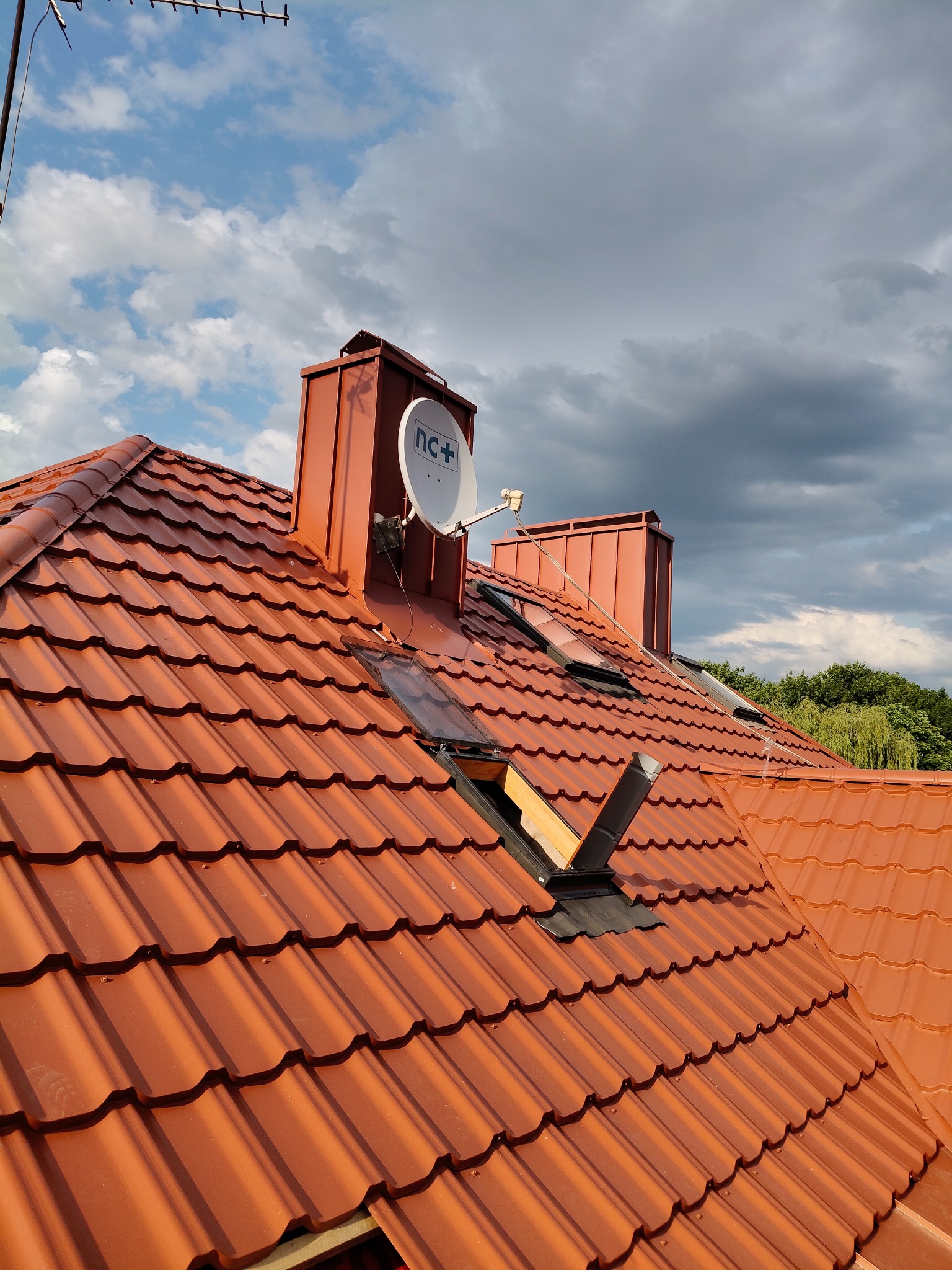 Czerwony dach z dachówki ceramicznej, komin z anteną satelitarną, okna dachowe. Częściowo wykonane prace dekarskie, widoczne elementy konstrukcji dachu. Błękitne niebo z chmurami.