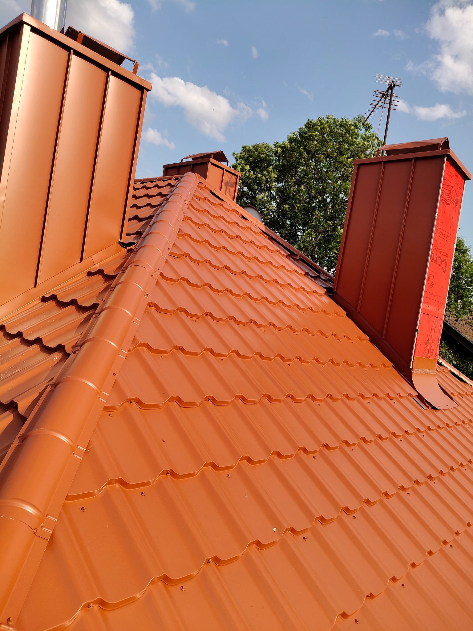 Nowy, miedziany dach z blachy trapezowej z kominami i anteną na tle błękitnego nieba z chmurami, zieleń drzew w tle. Widok z góry.
