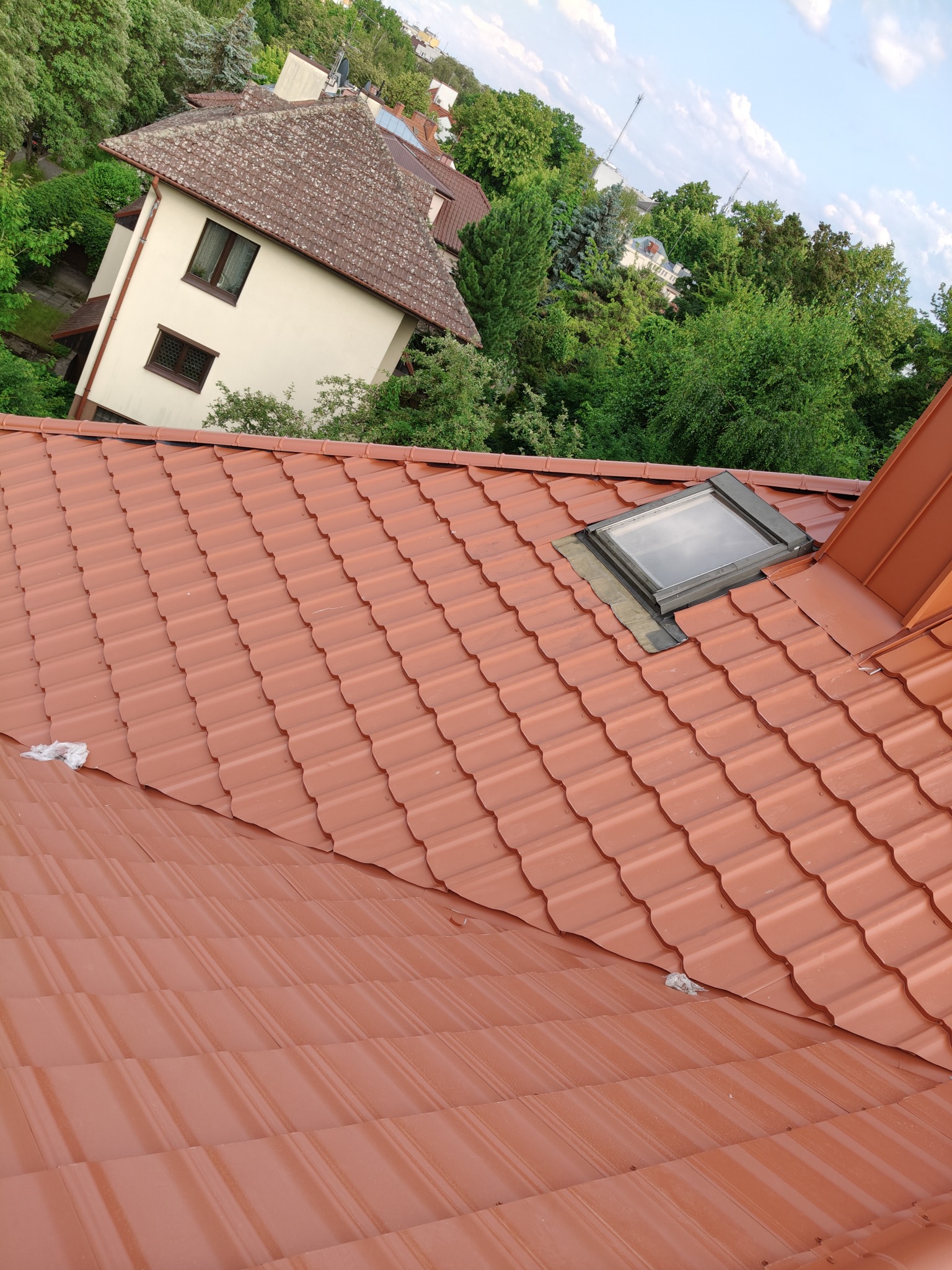 Czerwona dachówka ceramiczna z oknem dachowym na tle zieleni i budynku. Ujęcie z góry, podkreślające detale pokrycia.