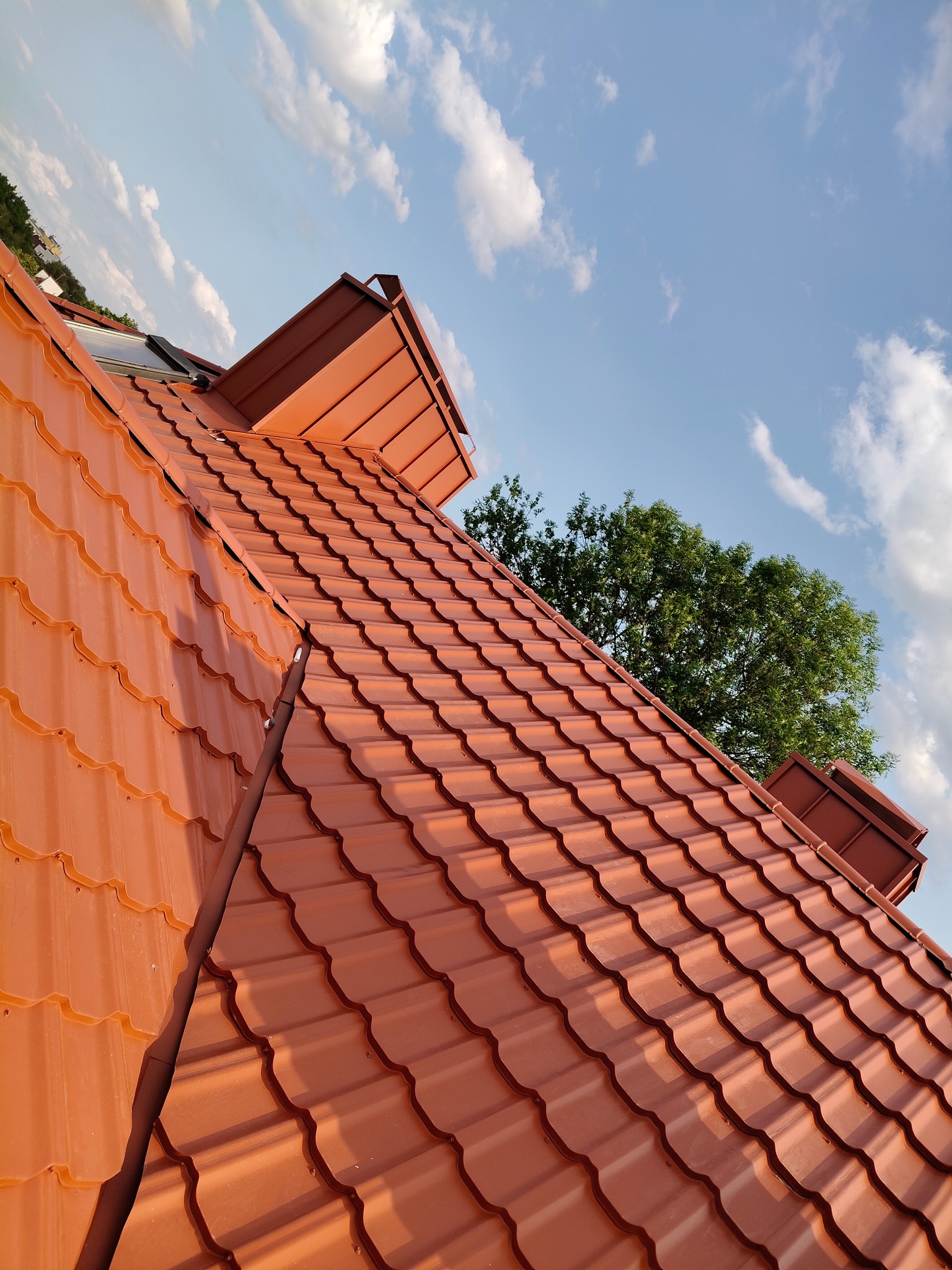 Czerwony dach z blachodachówki modułowej, widok z dołu w kierunku nieba z chmurami i fragmentem drzewa. Widoczne kominki wentylacyjne i okno dachowe.