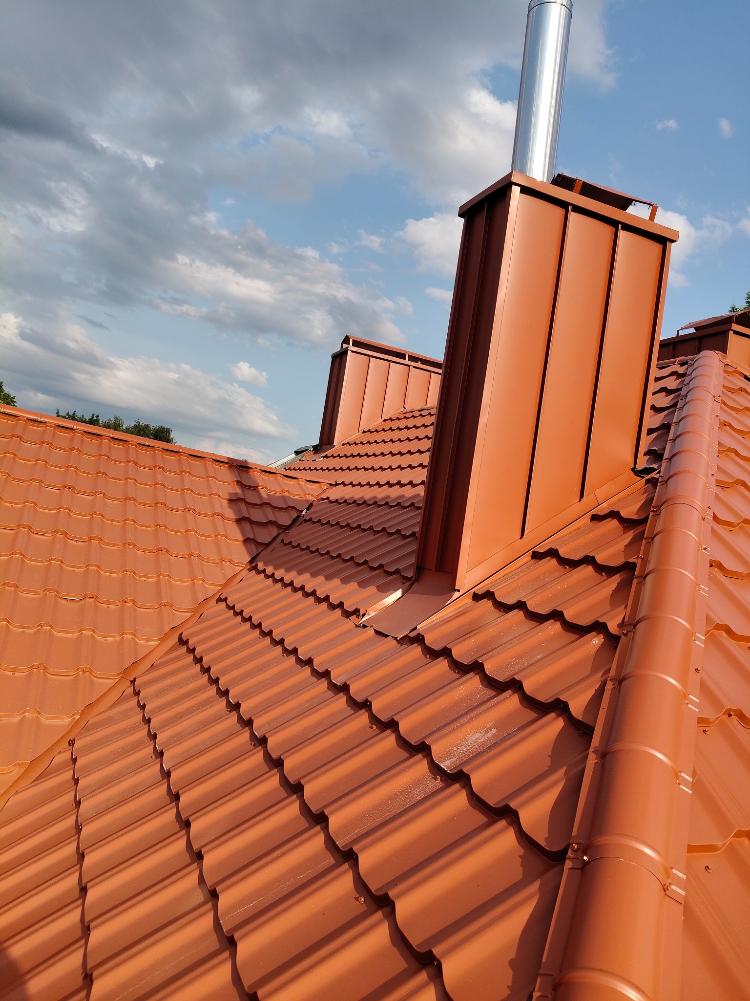 Nowy, pomarańczowy dach z blachodachówki trapezowej i komin z obróbką blacharską. Ujęcie z dołu, widoczne chmury na niebie.