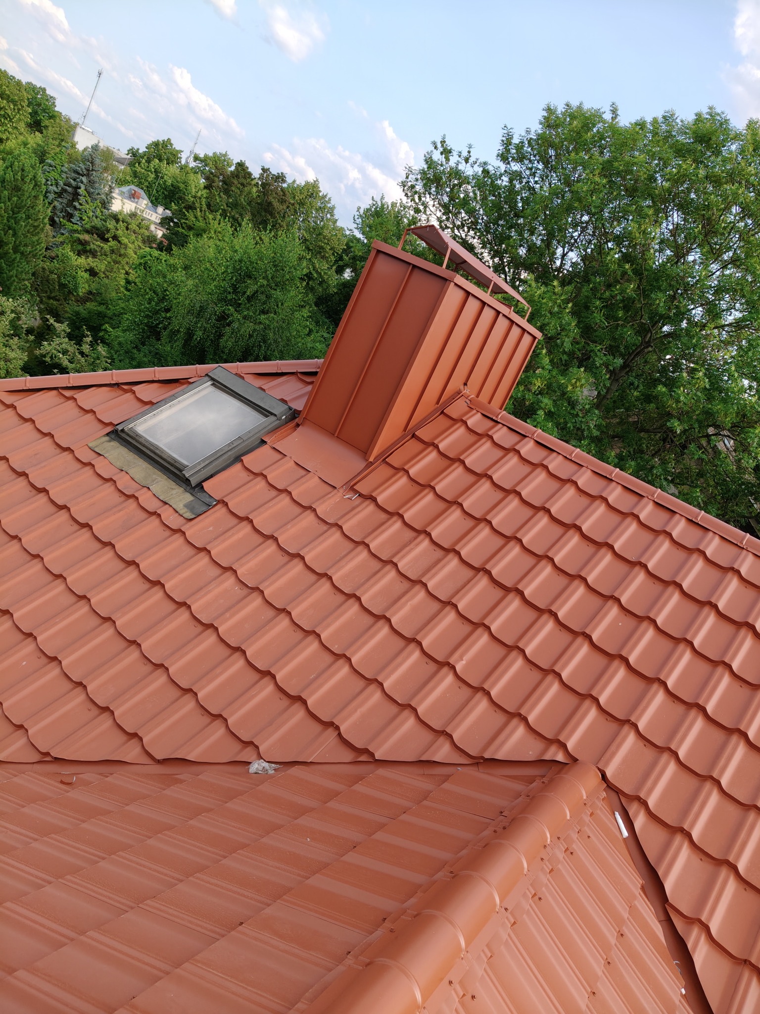 Czerwony dach z blachodachówki z oknem dachowym i kominem wentylacyjnym na tle zieleni. Widok z góry, delikatne zachmurzenie.