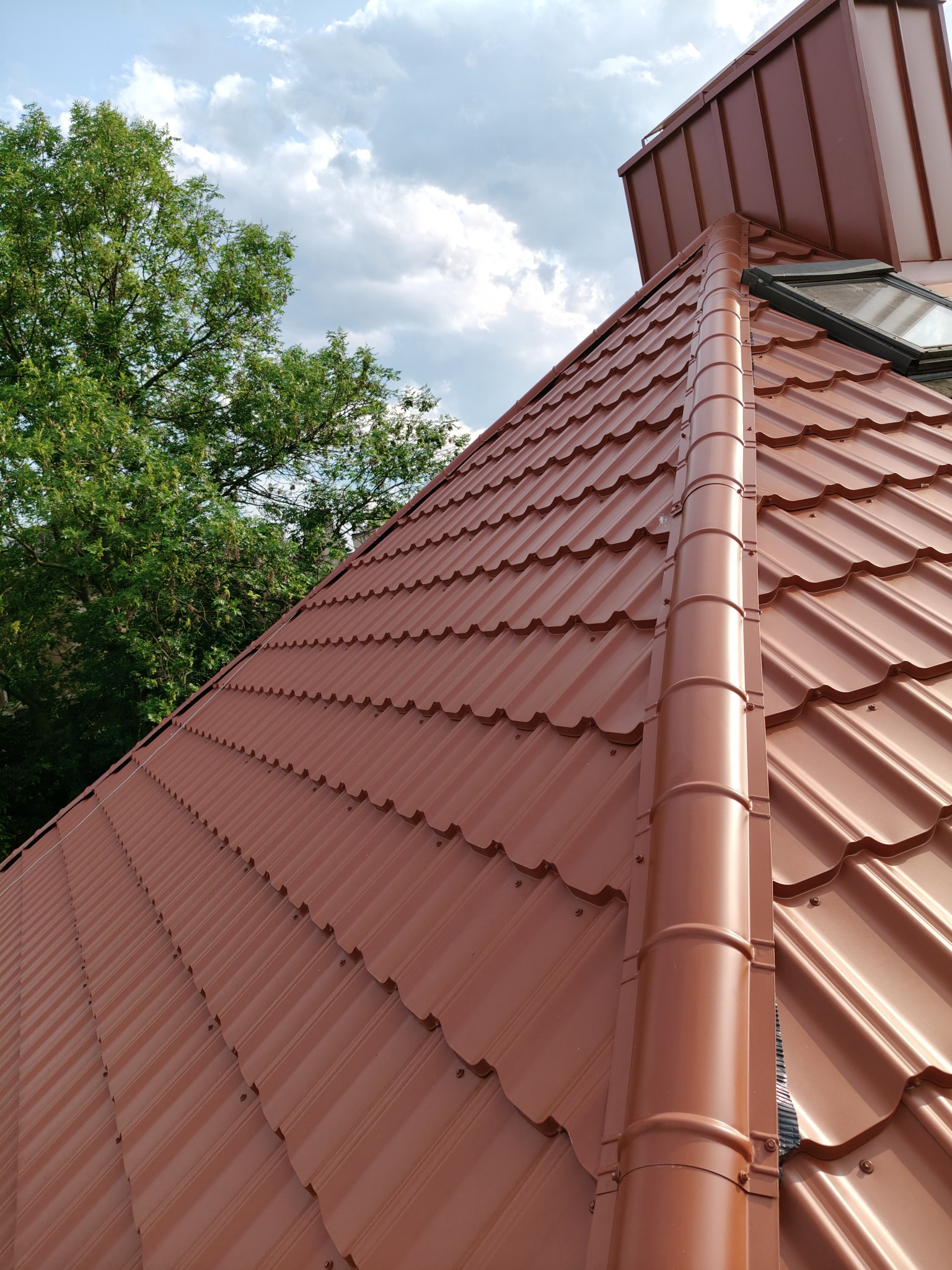 Nowy, brązowy dach z blachodachówki modułowej, widoczny z dołu pod kątem, z oknem dachowym i fragmentem drzewa na tle pochmurnego nieba.