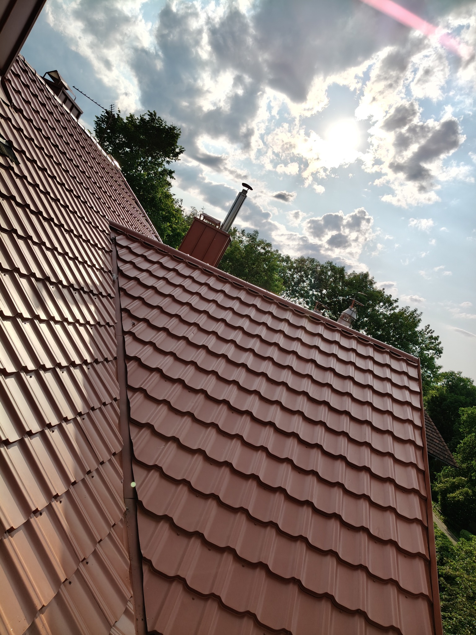 Nowy, brązowy dach z blachodachówki modułowej, z kominem i anteną, na tle błękitnego nieba z chmurami i słońcem. Widok z dołu, lekko pod kątem.
