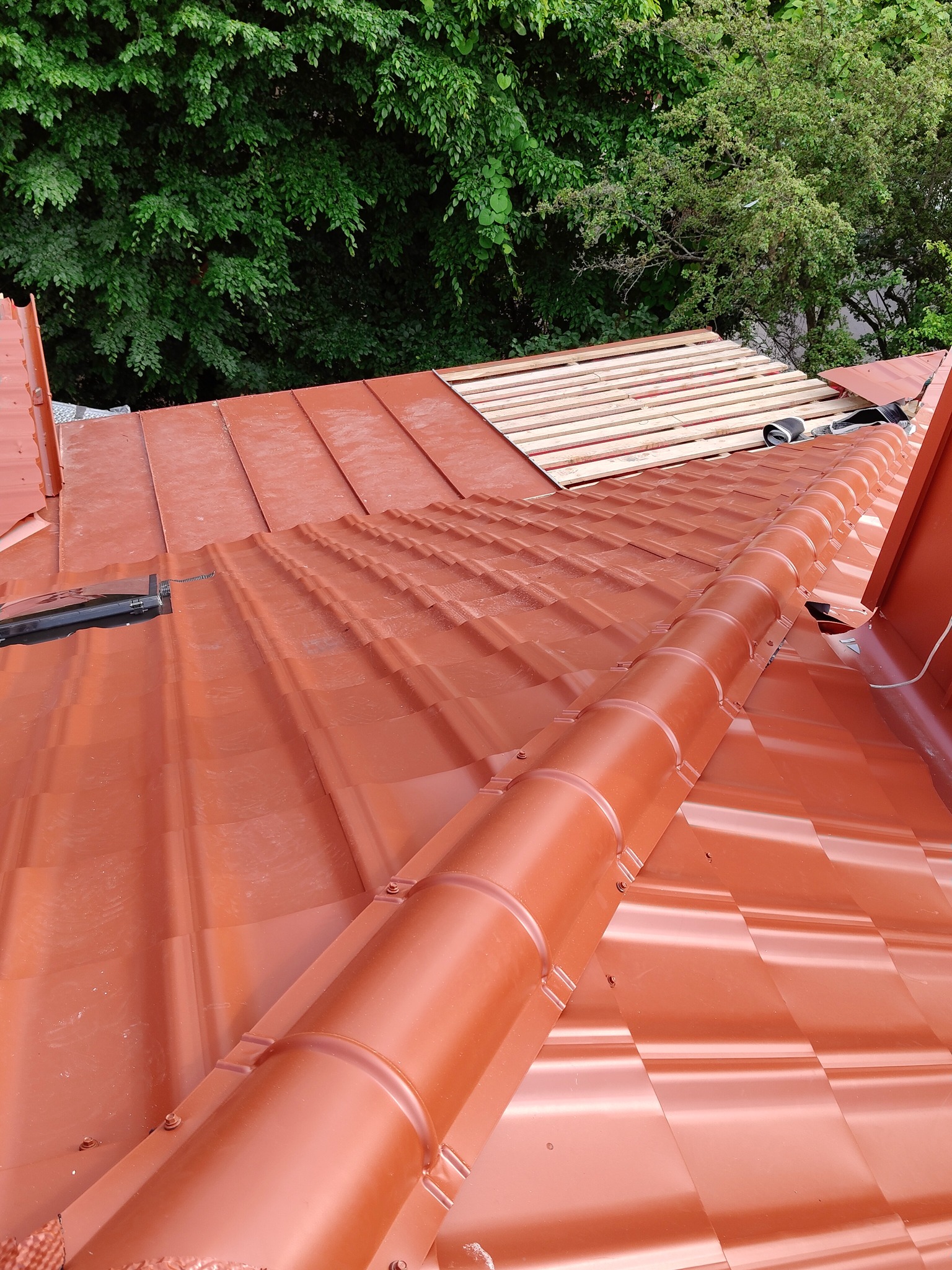 Czerwony dach z blachodachówki modułowej w trakcie montażu, widoczne łaty drewniane i elementy mocujące, tło stanowią zielone korony drzew. Ujęcie z góry.