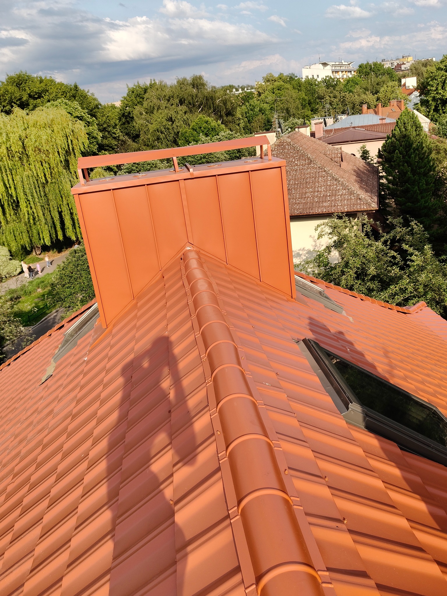 Nowy, pomarańczowy dach z dachówki ceramicznej, z oknem dachowym i kominem. Cień osoby na dachu. W tle zieleń drzew i zabudowa miejska.