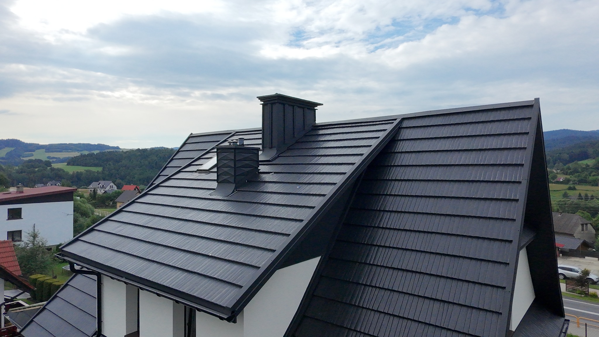 Czarny dach z blachy trapezowej z dwoma kominami na tle zielonych wzgórz i pochmurnego nieba. Widok z góry ukazuje elegancką, nowoczesną architekturę domu.