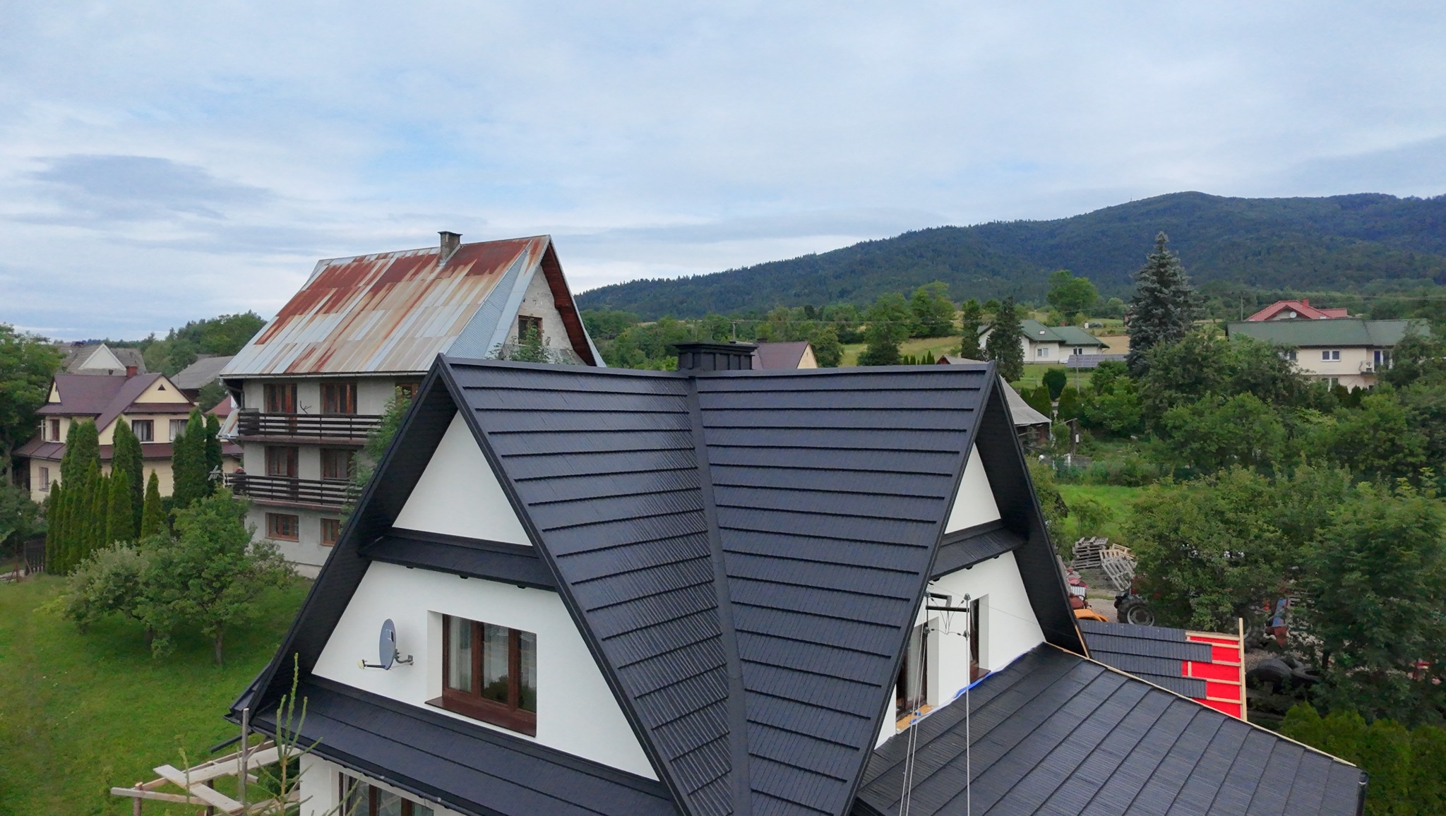 Czarny dach z blachy na białym domu, widok z góry na tle zieleni i gór. W tle dom z rdzawym dachem. Dachówka ułożona równo, symetrycznie.