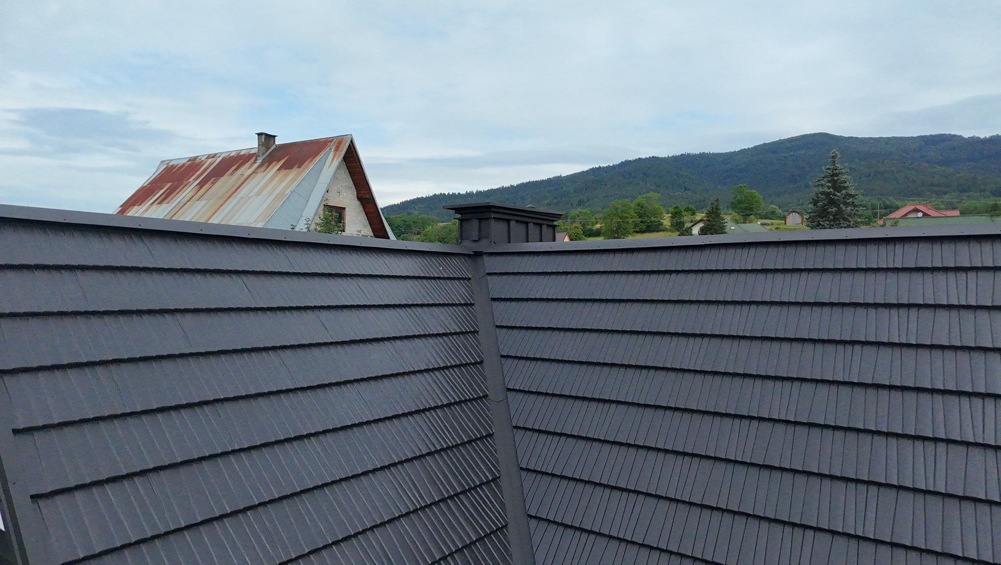 Nowy, grafitowy dach z dachówki bitumicznej, widok z góry, w tle budynek z rdzewiejącym dachem i zalesione wzgórza w pochmurny dzień.