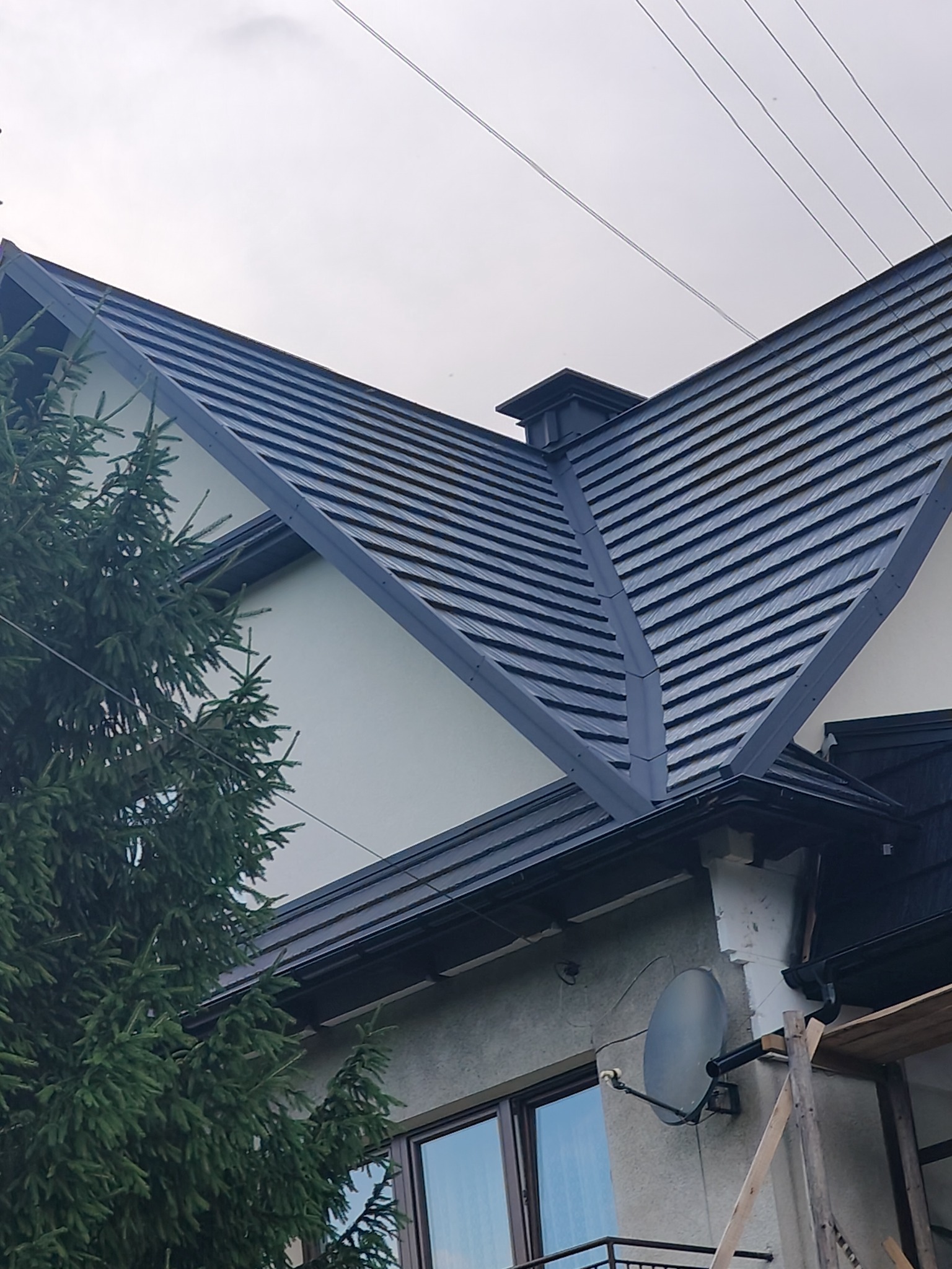 Szary dach z blachy na dwuspadowym budynku, widok z dołu, z fragmentem drzewa iglastego po lewej stronie. Widoczne okno i fragment rusztowania.