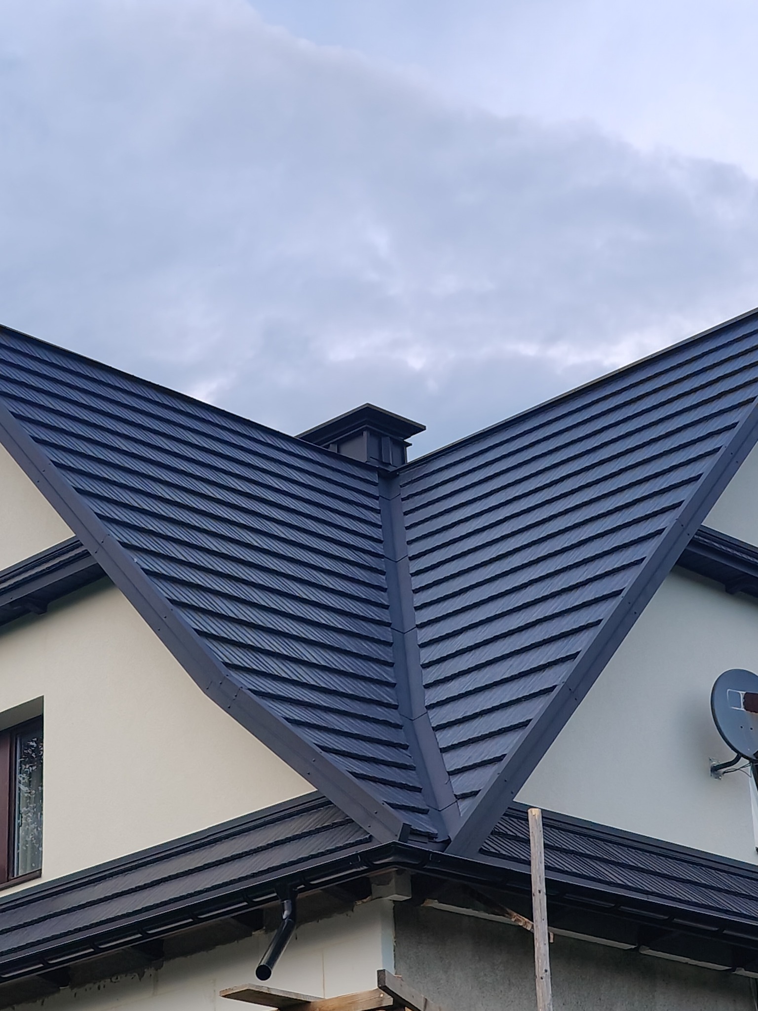 Ciemna dachówka modułowa na skośnym dachu domu jednorodzinnego. Widoczny komin i orynnowanie. Częściowo ocieplona ściana szczytowa. Pochmurne niebo w tle.