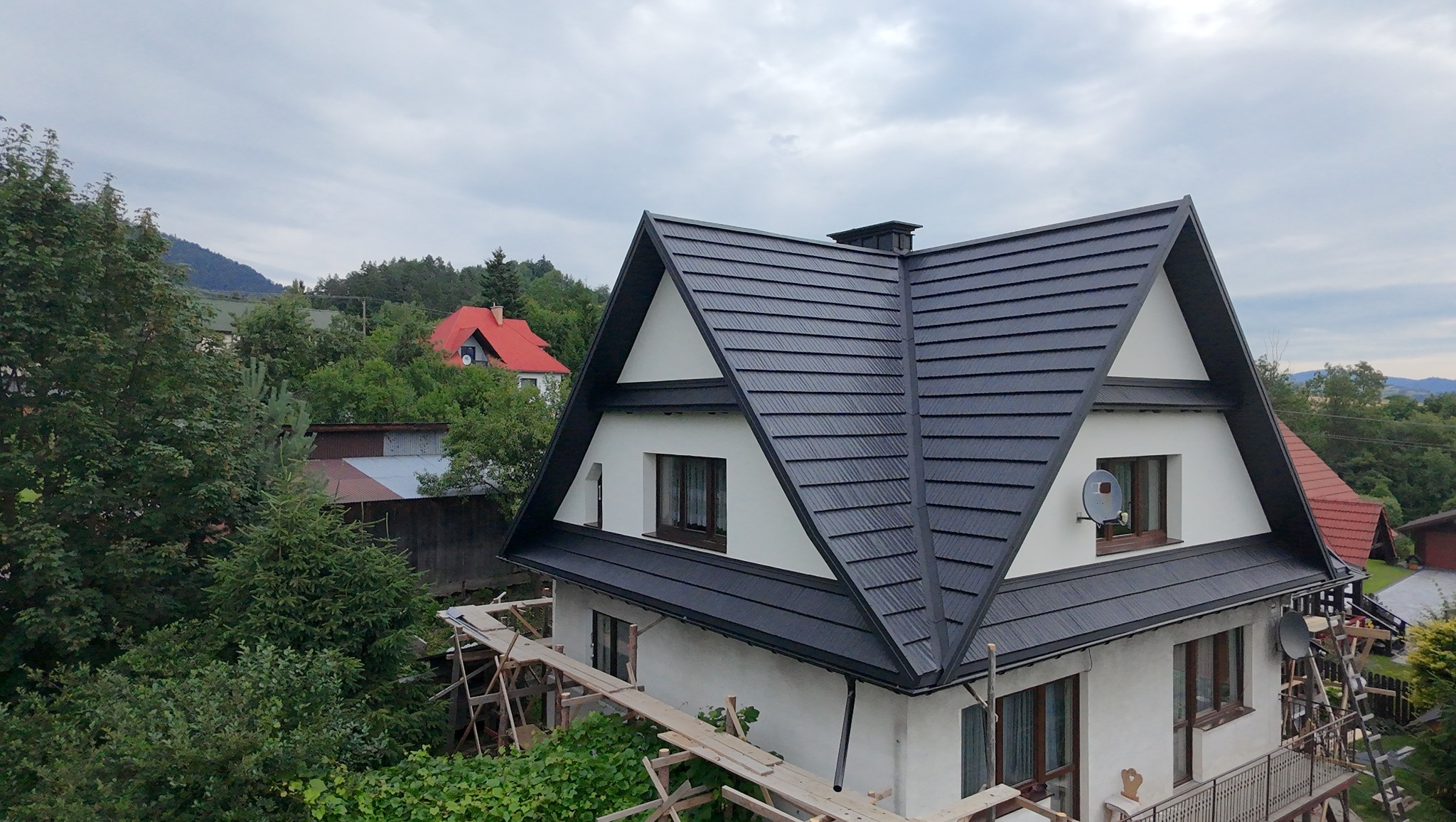Dom z czarnym dachem w kształcie litery A, widok z góry, otoczony zielenią. Widoczne rusztowania. W tle wzgórza i inne budynki.