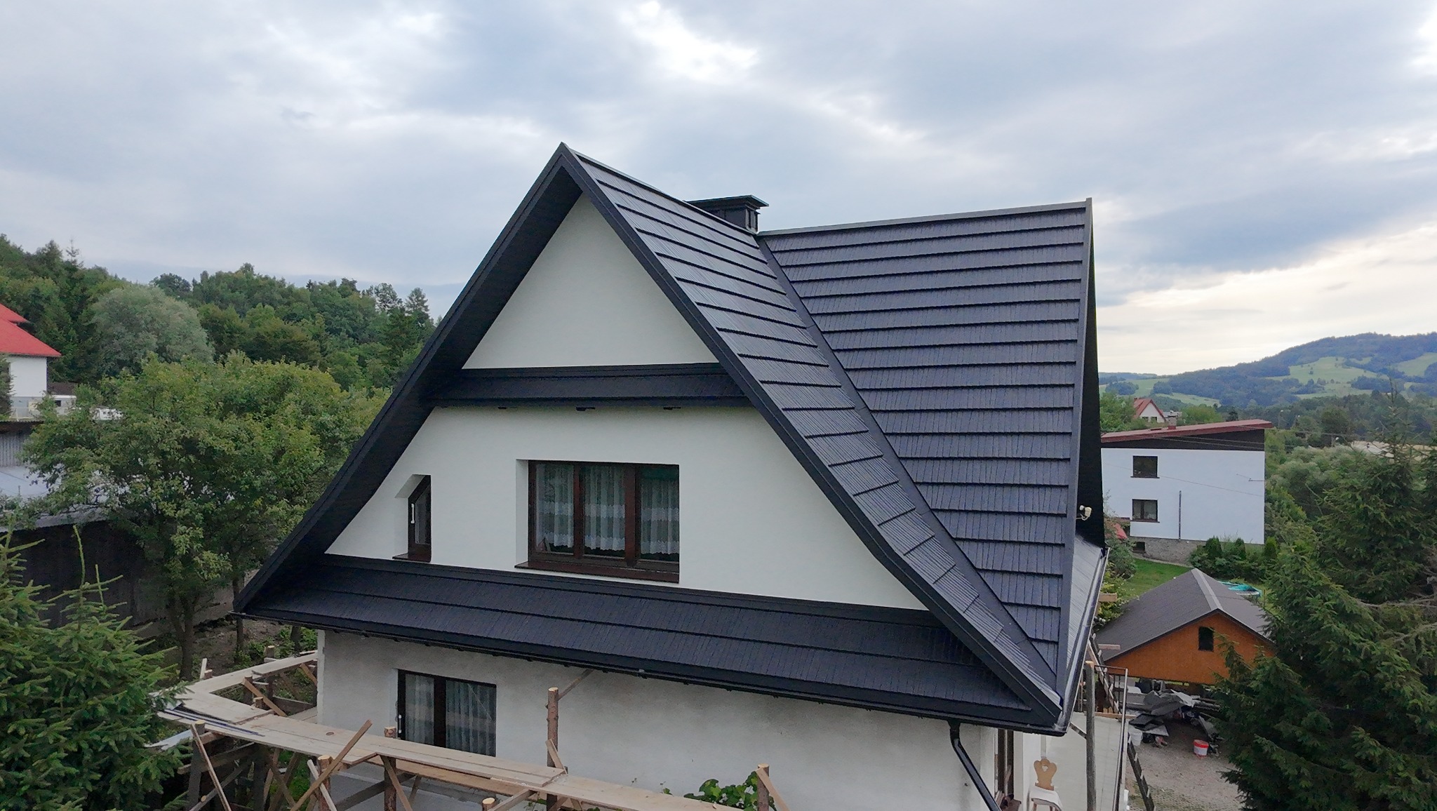 Dom jednorodzinny z czarnym dachem z blachy na rąbek stojący, widoczny komin i okna. W tle zieleń drzew i wzgórza. Częściowo widoczne rusztowanie.