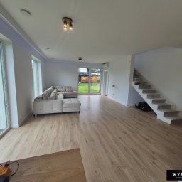 WYMALOWANI - Jasne, przestronne wnętrze po remoncie: panele podłogowe, pomalowane ściany na biało, surowe schody betonowe, sofa narożna i duże okna z widokiem na zieleń.