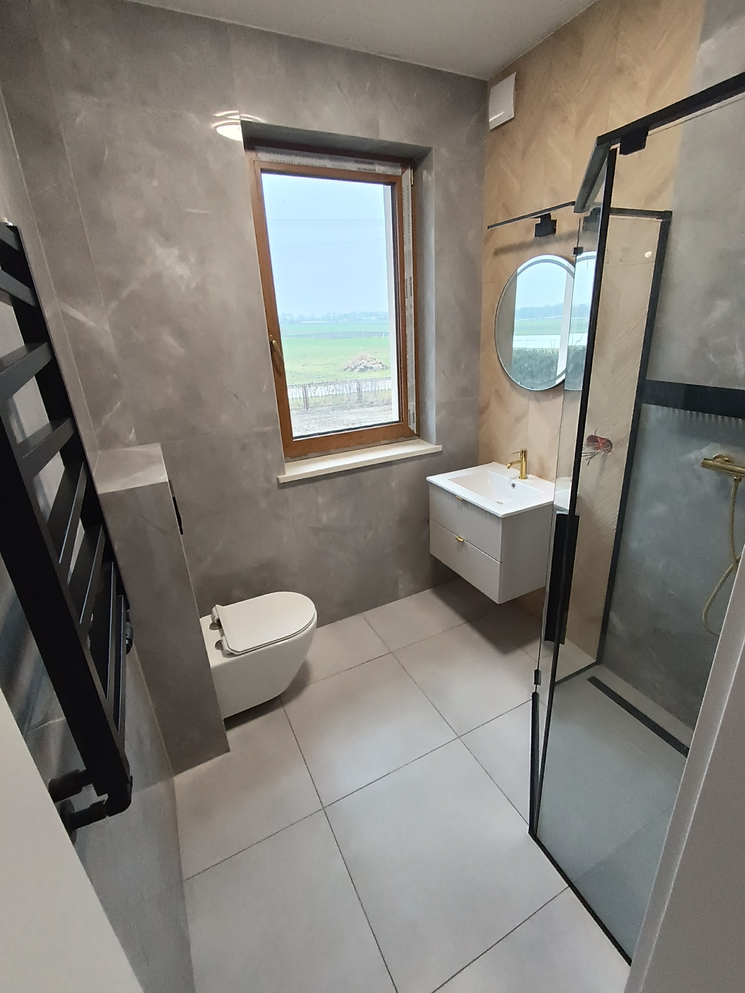 Nowoczesna łazienka z szarymi płytkami imitującymi marmur, drewnianym oknem z widokiem na zielone pole, okrągłym lustrem i złotą armaturą.