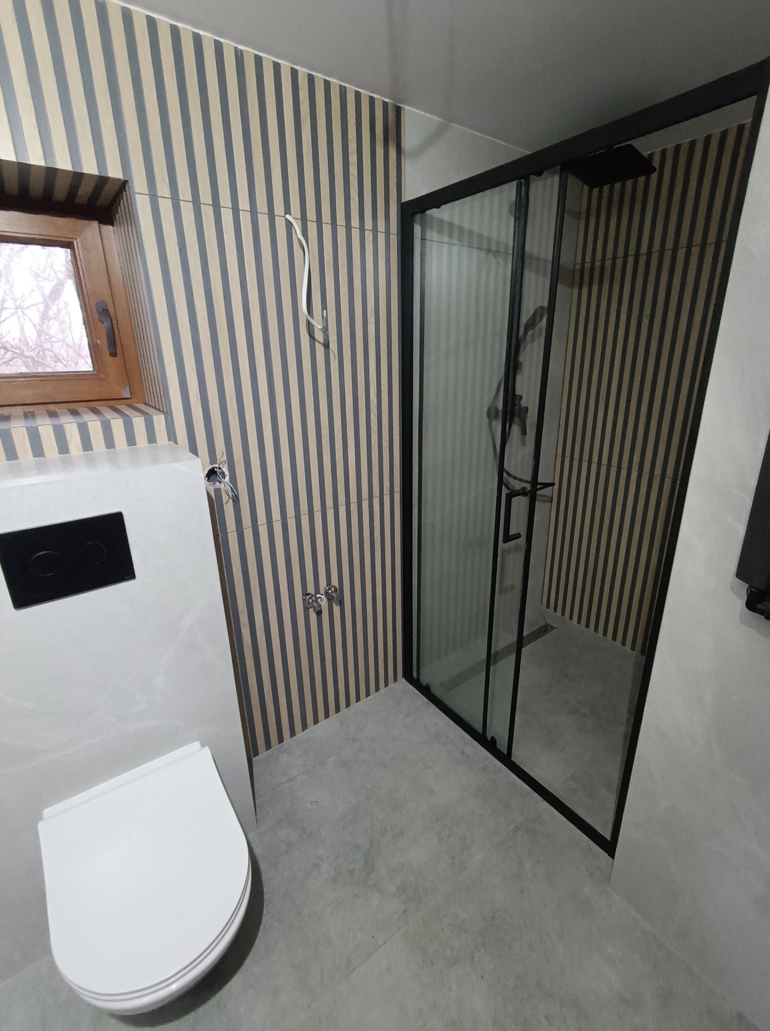 Nowoczesna łazienka z kabiną prysznicową typu walk-in z czarnymi ramami, wiszącą toaletą z czarnym przyciskiem spłukującym oraz ścianami wykończonymi w pionowe pasy beżu i szarości.